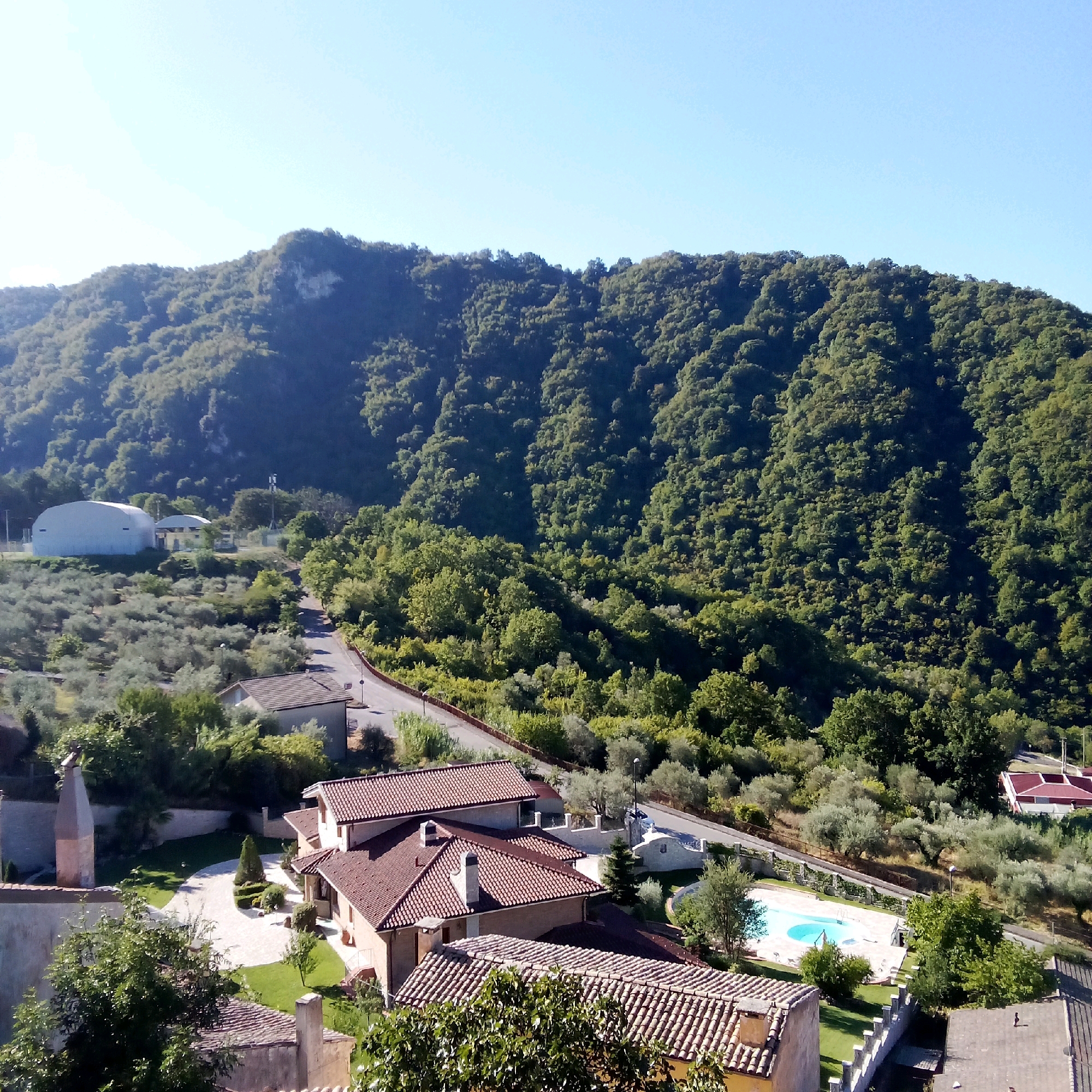 This is an aerial view of a rural Italian landscape, likely in or near Santomenna, Italy, on a clear day. The scene is dominated by a lush, green, tree-covered mountain that rises in the background. In the foreground, there are several buildings with red tile roofs, characteristic of Italian architecture. One building is a light-colored structure with a prominent chimney. Another appears to be a multi-story residential building. A swimming pool with blue water is visible to the right, surrounded by a paved area and greenery. A paved road winds through the scene, lined with trees and vegetation. There are also several olive trees scattered throughout the landscape. In the middle ground, a large, white, dome-shaped structure, possibly a warehouse or industrial building, is visible. The overall impression is one of a tranquil, scenic countryside setting with some modern infrastructure. There are no people visible in the image.