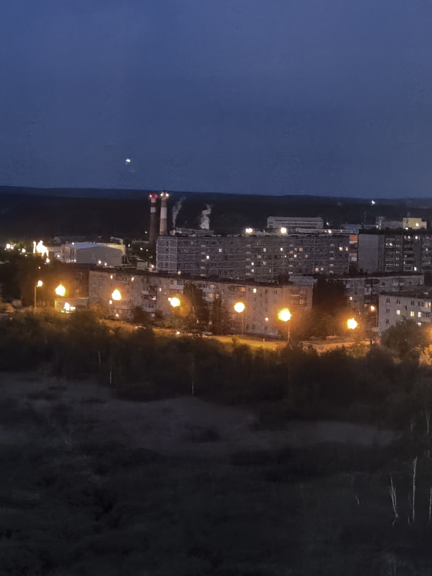This is a nighttime, medium-distance shot of a residential area with some industrial structures in the background. In the foreground, there is a dark, wooded area with some visible trees. Beyond the trees, apartment buildings are visible, illuminated by lights from within the windows and by streetlights. The streetlights are a warm, orange color. In the background, there are industrial buildings, one of which has two tall smokestacks emitting white smoke. The sky is a dark blue, indicative of twilight or night, with a faint, bright object visible in the upper left portion of the sky.