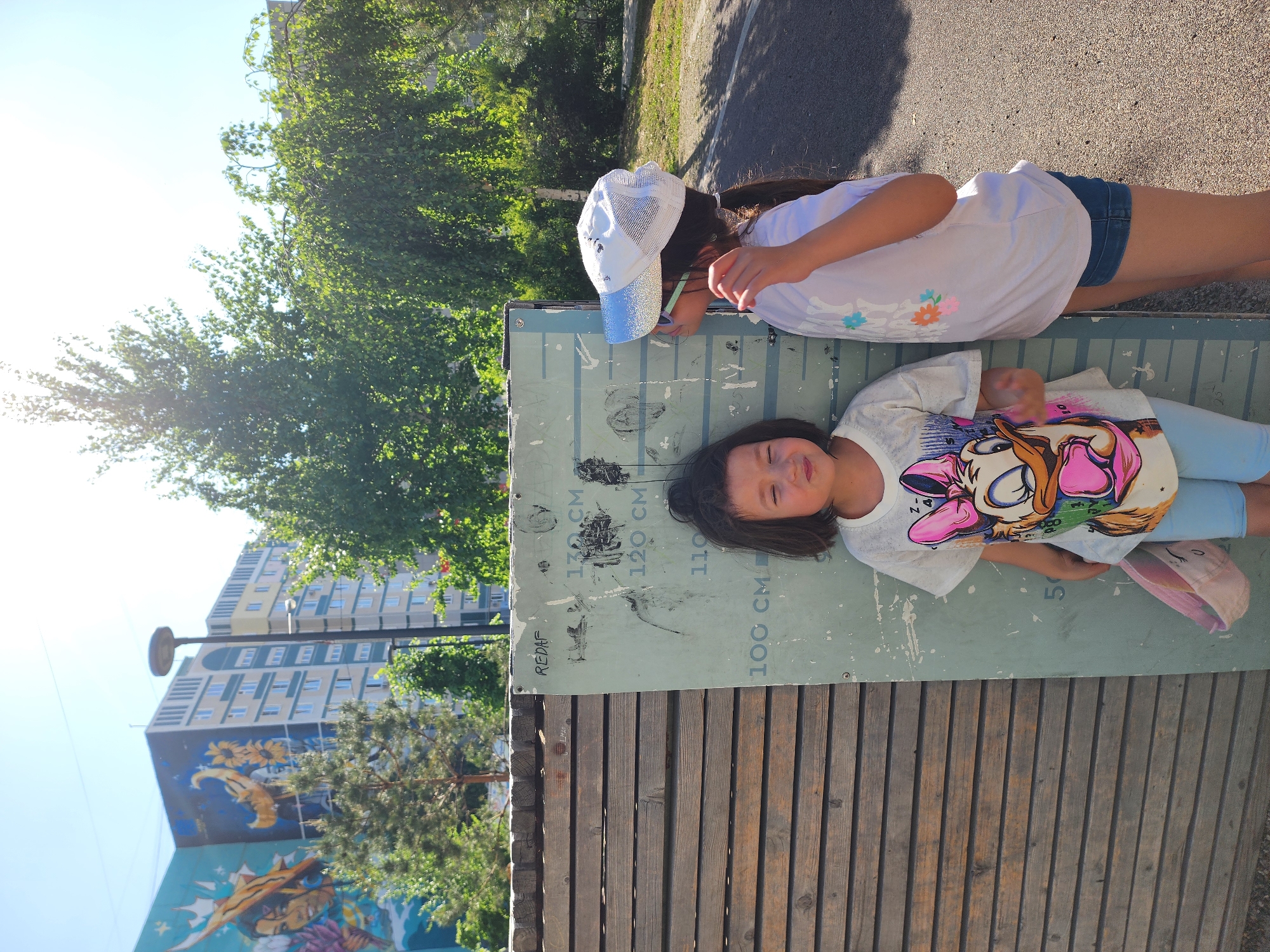 Two girls are lying on a wooden bench with a height chart attached to it. The younger girl is lying on the chart with her head at the 120 cm mark. She is wearing a t-shirt with a cartoon graphic and light blue leggings. The older girl is wearing a white t-shirt, denim shorts, and a white and blue baseball cap. She is standing next to the bench, looking at the height chart. The bench is located in a park with trees and a building with a mural in the background. The mural is colorful and depicts various sea creatures. The location is Kazan, Russia.