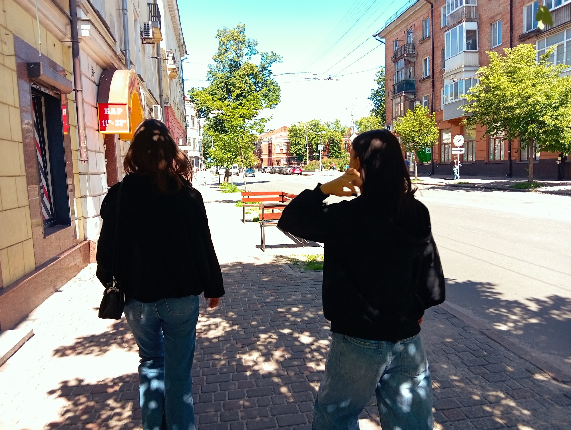The image depicts an outdoor urban scene on a bright, sunny day, likely in Poltava, Ukraine. Two young women, viewed from behind, are walking away from the camera on a paved sidewalk. Both are dressed in black long-sleeved tops, possibly hoodies or sweatshirts, and blue jeans. The woman on the left carries a small black shoulder bag. The woman on the right has her hand raised near her head, perhaps adjusting her hair or shading her eyes.
Buildings line both sides of the street. On the left, a light-colored building features a prominent red and yellow sign that reads "БАР" (BAR) and "11-23," indicating a bar open from 11 AM to 11 PM. Further down the street, on the right, a multi-story building constructed of reddish-brown brick with several balconies is visible.
Green trees are scattered along the street and between buildings, casting clear, dappled shadows onto the sidewalk, characteristic of bright sunlight. A red-painted wooden bench is situated in the midground. The street itself can be seen extending into the distance, with parked cars and some vehicles in motion. Overhead utility wires crisscross against a clear, light blue sky. The scene conveys a sense of a casual walk on a pleasant day in a city environment.