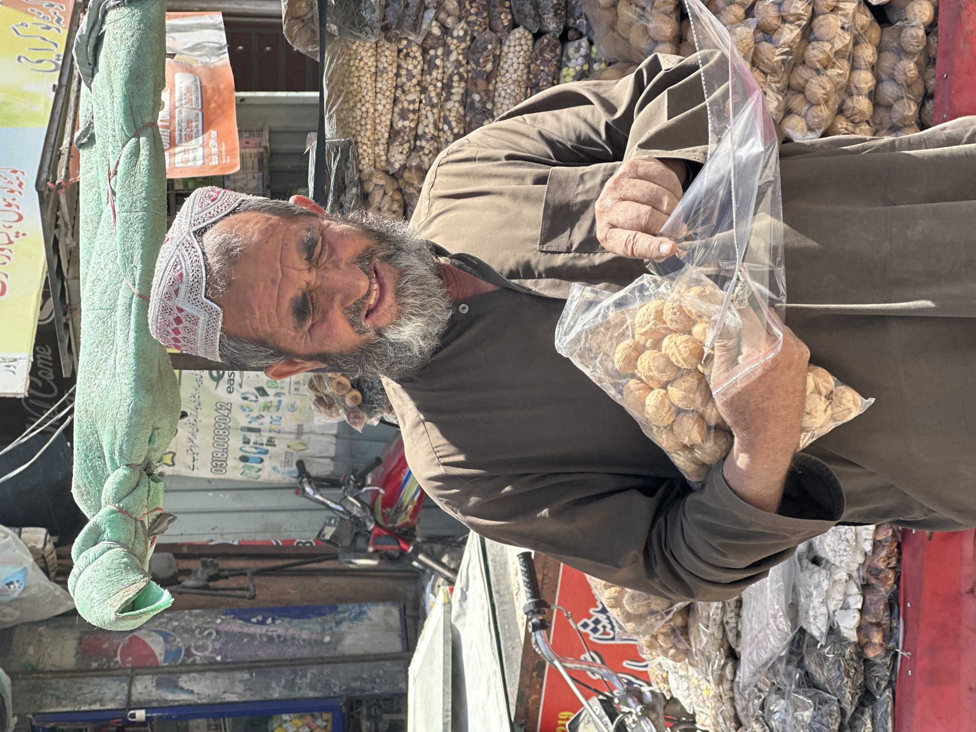 An outdoor market scene captures a smiling man, likely a vendor, looking directly at the camera. He has a grey beard and wears a brown long-sleeved tunic and a white skullcap adorned with red and pink embroidery. In his hands, he holds two clear plastic bags, each filled with whole walnuts in their shells, presenting them to the viewer.
Behind him, a wide array of similar clear plastic bags filled with various nuts, dried fruits, and seeds (including chickpeas, walnuts, and dried figs) are neatly displayed on shelves or stalls, indicating a bustling marketplace. The setting appears to be a sunny daytime street scene, with bright light and visible shadows.
Numerous commercial signs in the background are written in Urdu script. Visible text includes "زی پیسہ اور بیٹری جاکر تو ہیڈ فون اور عطر دستیاب ہے" (Easy money and battery charger, headphones and perfume available), a phone number "0318.0089042", and the city name "کوئٹہ" (Quetta). Other partial texts like "GB Dota", "Booker Snow", and a Pepsi logo are also discernible. A green, rolled-up fabric hangs vertically on the left side, and parts of a red motorcycle are visible in the mid-ground. The man's friendly expression adds to the lively atmosphere of the market.