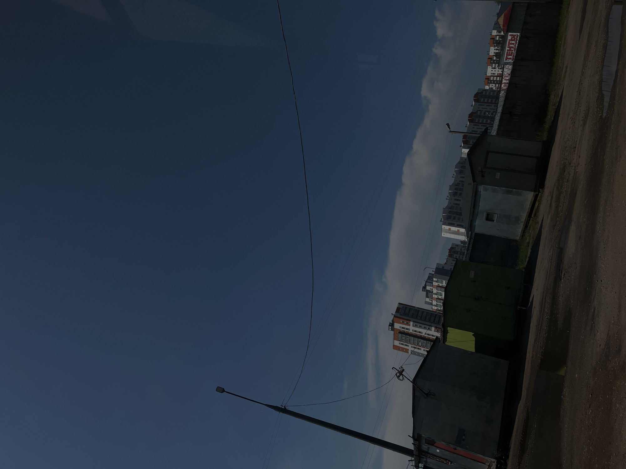 This image shows an outdoor scene featuring a row of garages and apartment buildings under a bright blue sky with some clouds. The perspective is from ground level, looking upwards and slightly to the right. Several garages, some painted green and others a dark grey, are in the foreground. A street lamp is visible on the left, with power lines stretching across the sky. In the background, a series of multi-story apartment buildings with white, orange, and grey facades are clustered together. The overall setting appears to be an urban or semi-urban residential area. The time of day seems to be daytime, given the bright lighting and blue sky. There are puddles on the ground near the garages, suggesting recent rain or a damp area. No people are visible in the image. The only visible text is on a banner on a fence behind the garages, but it is too blurry to read. The scene evokes a sense of everyday urban life in Vitsebsk, Belarus.