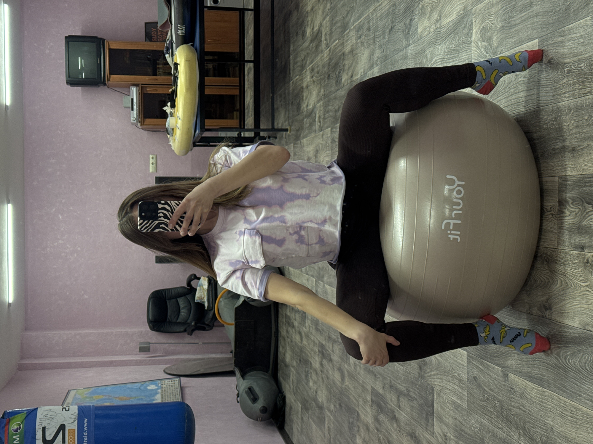 The image shows a woman in a room in Прикамский, Russia. She is sitting on a large beige exercise ball wearing dark leggings and a light purple and white t-shirt. Her face is obscured by her phone, which has a zebra-striped case. She is wearing socks with a banana print. The room has light purple walls and a gray wood-look floor. There is a black office chair, a blue cylinder, a weight bag, and various pieces of furniture including a TV and wooden shelving in the background.