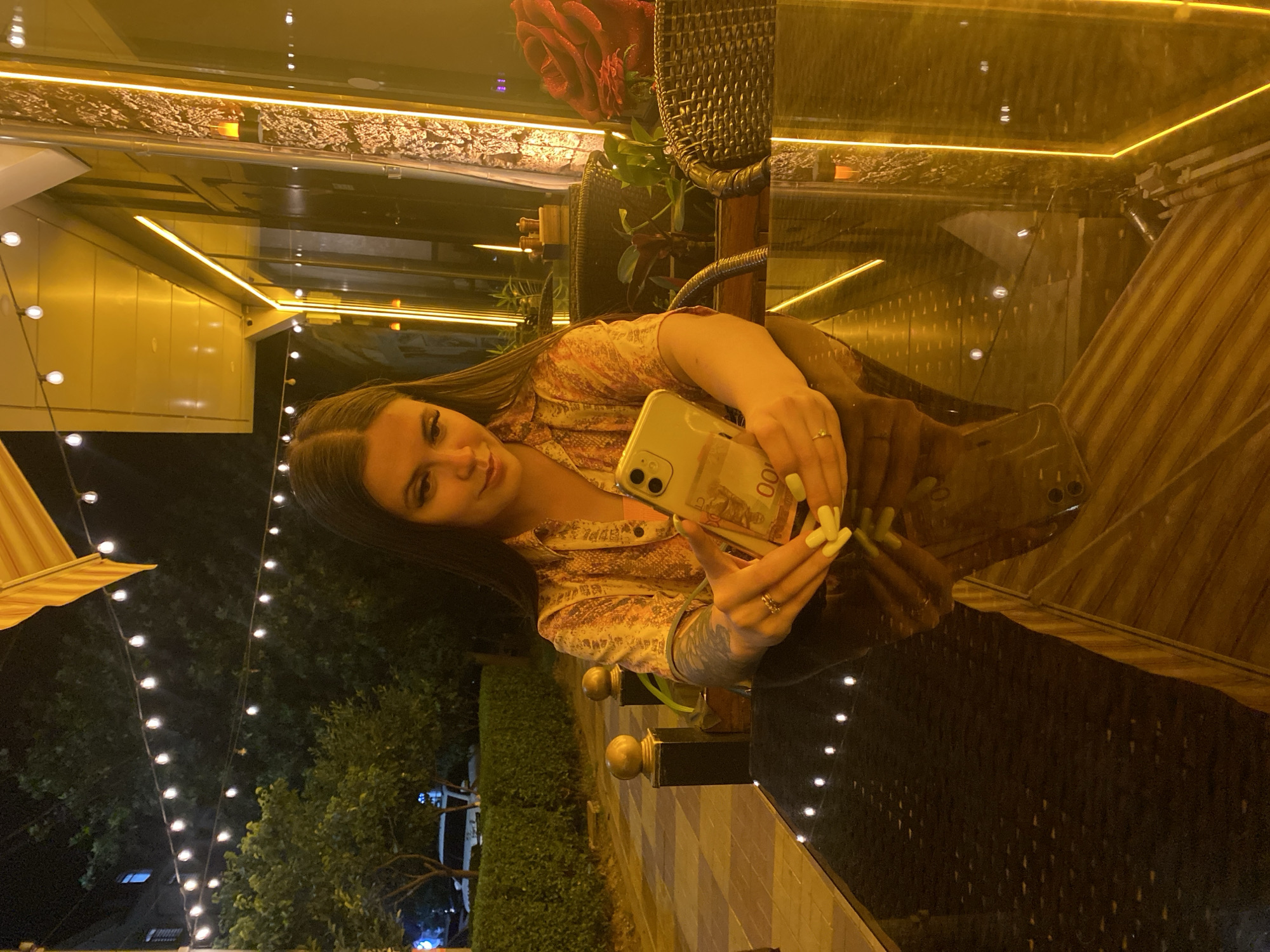 A young woman with long dark hair is sitting at a table outside a restaurant at night. She is wearing a patterned shirt and holding a smartphone. The table is made of dark, reflective material, and string lights hang overhead. In the background, there are trees, hedges, and the illuminated facade of the restaurant. The scene appears to be in Derbent, Russia, indicated by the architecture and flora.