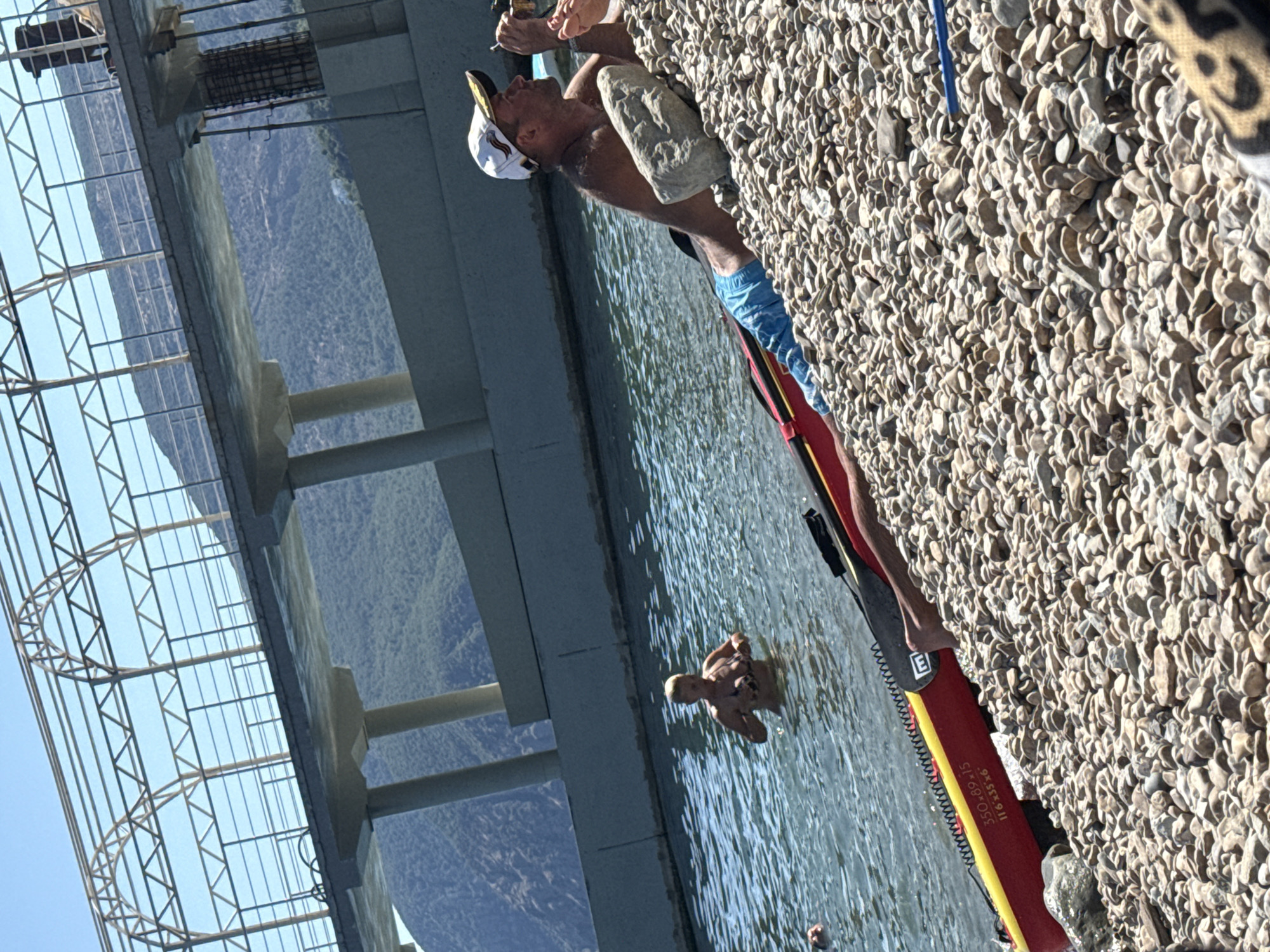 A man wearing a white baseball cap and blue shorts reclines on a pebble beach next to a red and yellow inflatable kayak. He is holding a cigarette and appears to be looking at something off-camera. In the background, a woman with blonde hair stands in the water, wearing a dark bikini. The scene is set at the waterfront in Utios, Ukraine, with a large concrete structure and a bridge visible behind them. Mountains can be seen in the distance.