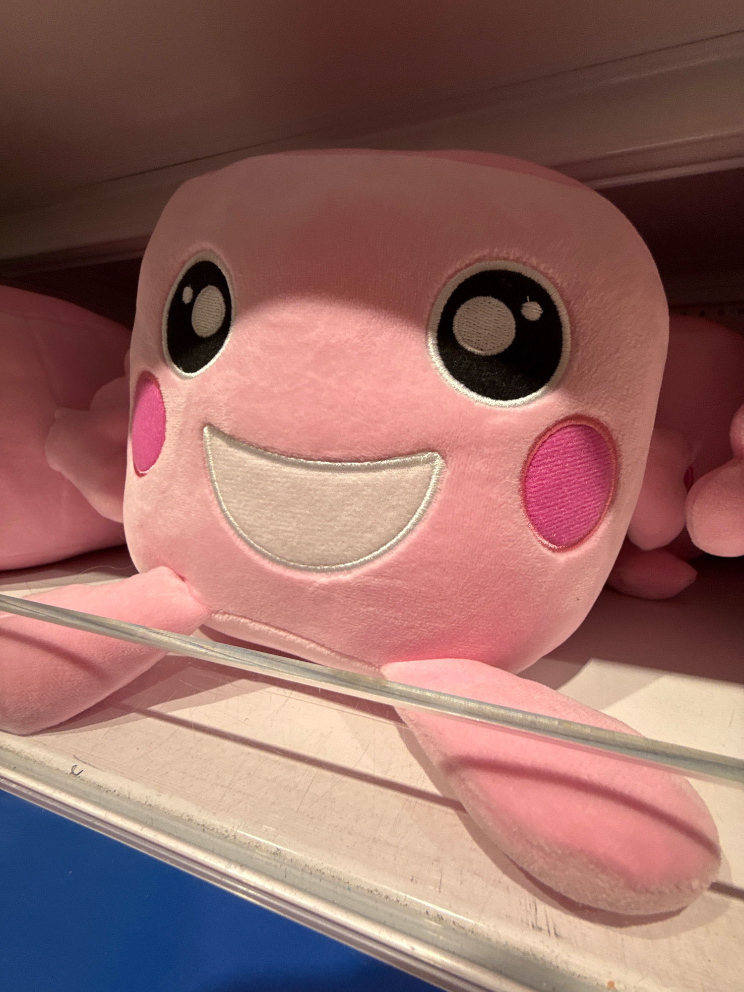 A close-up, low-angle shot shows a bright pink plush toy with a large, smiling face. The toy has big, black, round eyes with white pupils and eyelashes. Two pink circles are embroidered on its cheeks. Its mouth is a wide, white, stitched smile. The toy is resting on a white shelf in what appears to be a retail environment, possibly a toy store. The shelf has a clear plastic barrier running horizontally across it, in front of the toy. The background is out of focus, with hints of a dark ceiling and other pink plush toys. This image was taken in Wolfsburg, Germany.