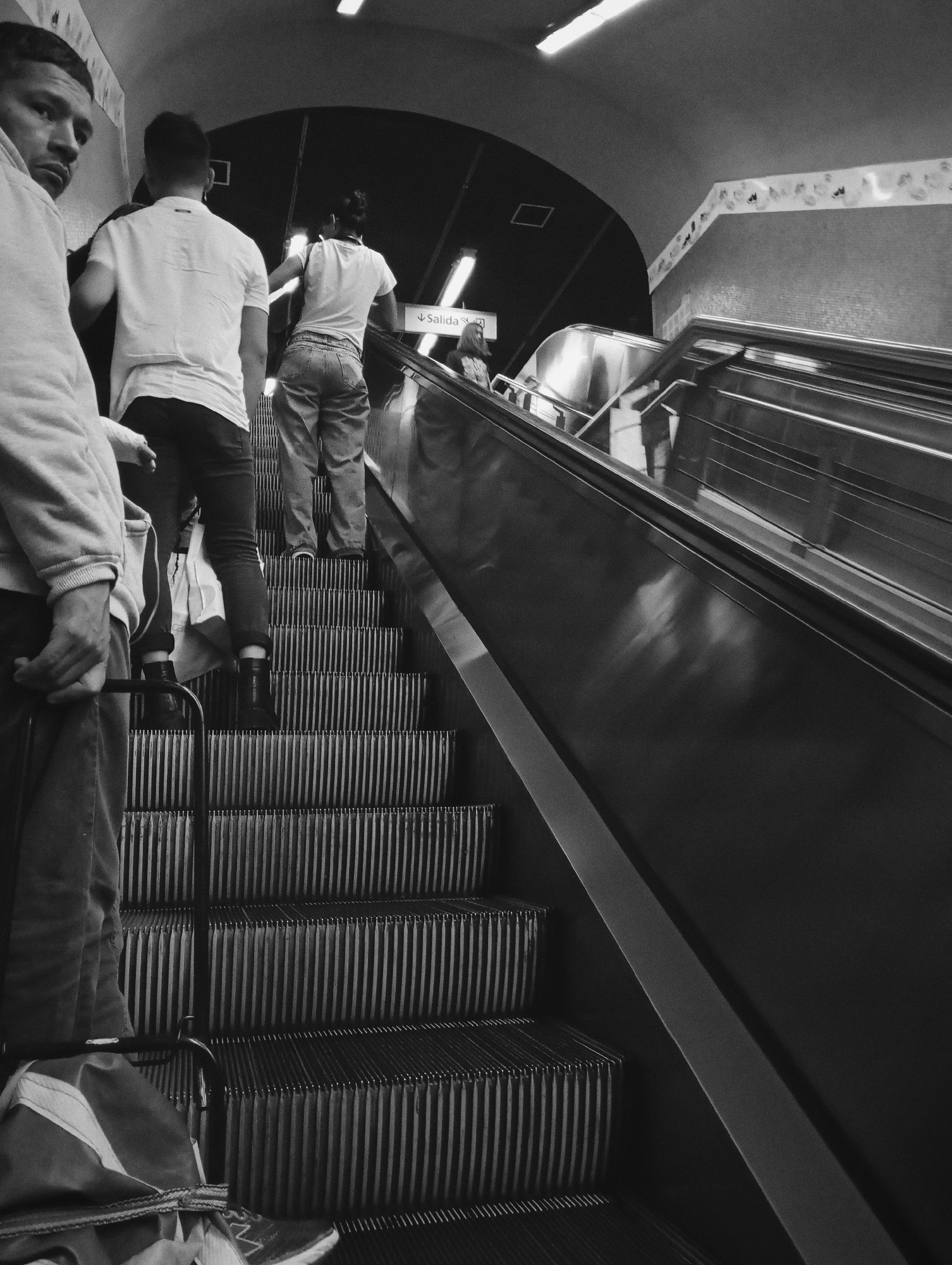 The black and white image captures a group of people ascending an escalator in an underground public transit station, identified as being in Buenos Aires, Argentina, by a visible "Salida" (Exit) sign.
In the foreground, a man is partially visible on the left, looking directly towards the viewer. He wears a light-colored hooded garment, dark trousers, and a sneaker with visible stripes. His right hand is positioned towards the bottom left of the frame.
Immediately behind him, a person is seen from the rear, ascending the escalator. This individual wears a white short-sleeved t-shirt, dark trousers, and dark ankle boots, holding a white bag in their left hand. Further up the escalator, another person is visible from behind, wearing a light-colored t-shirt and light-colored jeans. A fourth person's partial reflection or figure is faintly visible on the right escalator panel in the mid-ground.
The escalator features metallic, ribbed steps and a dark, possibly rubber, handrail on the right side. The station environment includes an arched ceiling with visible rectangular fluorescent light fixtures. A patterned decorative band runs along the upper section of the right wall, which appears to have a textured or tiled surface below. A clear "Salida" sign, displaying white text and an upward-pointing arrow on a dark background, is affixed to the wall above the escalator, indicating the direction of the exit.
