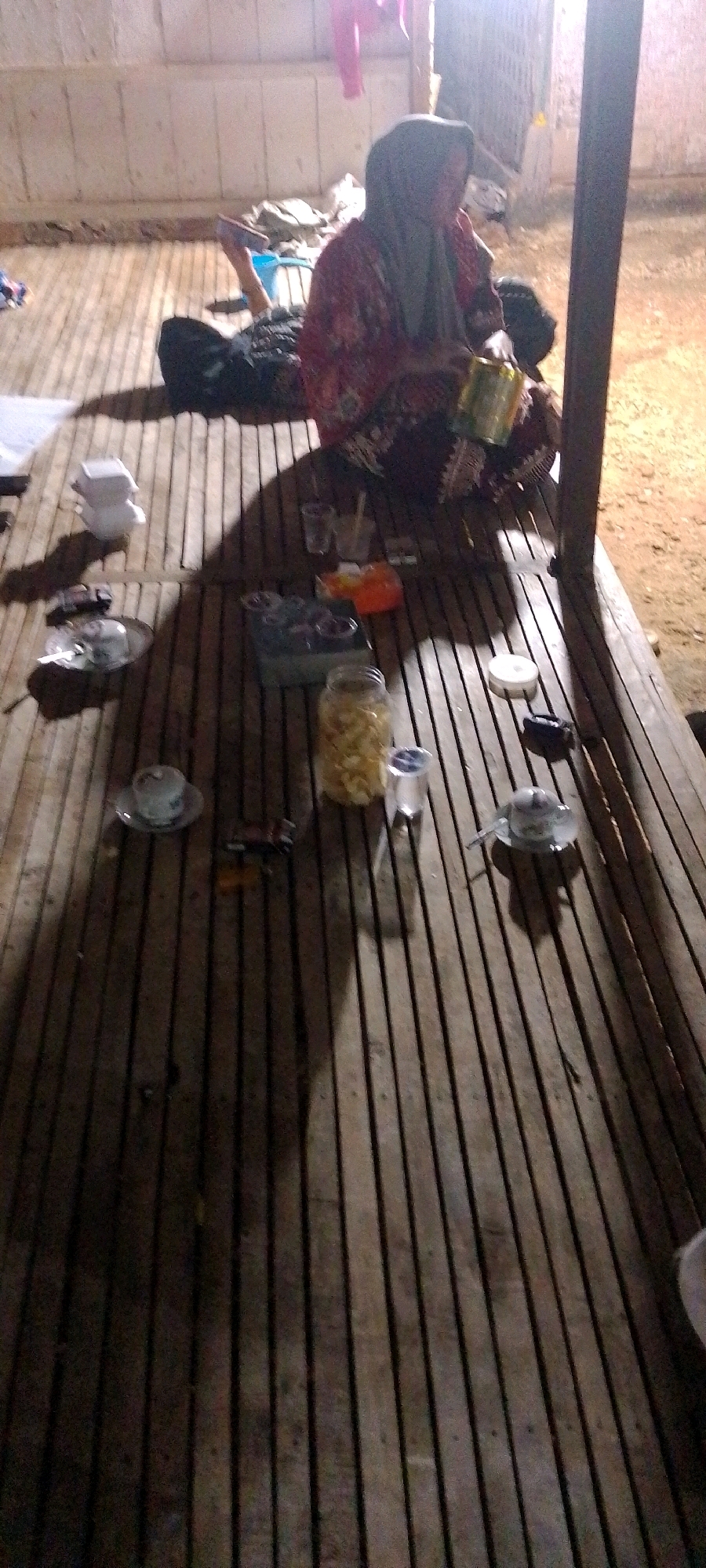 In a dimly lit, open-air structure with a bamboo slatted floor and wooden walls, two individuals are present. On the right, a woman wearing a hijab and a patterned robe is seated, holding a metal can. Her left arm is bent, and her hand is near her face. To the left of the seated woman, a second individual, whose face is obscured, lies on their back on the floor. This person is wearing dark clothing. Scattered on the bamboo floor are several objects, including cups, a jar filled with a light-colored crunchy food item, and a stack of white disposable containers. The environment appears to be rustic, possibly a temporary shelter or an outdoor seating area in a rural or less developed part of Pangereman, Indonesia. The lighting suggests it is either early morning or late evening.