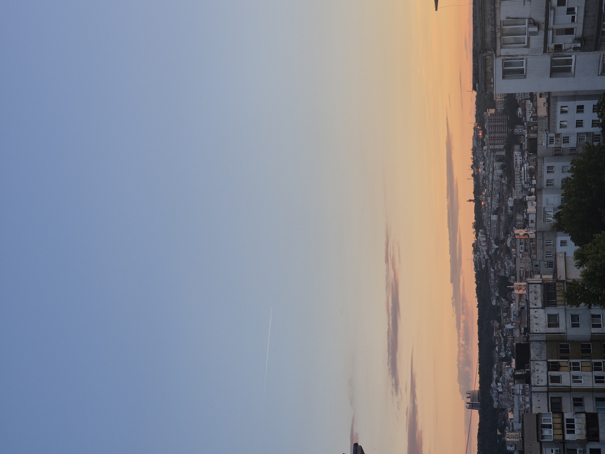 The image shows a cityscape at sunset. The sky is a gradient of blue to orange, with some wispy pink clouds. A contrail from an airplane is visible in the upper part of the sky. In the foreground, there are several multi-story apartment buildings. The buildings are concrete and appear somewhat dated. In the background, there is a dense urban area with more buildings, some of which are taller and appear more modern. A large, distinctive building with a rounded top is visible on a hill in the far background. Trees are present in the foreground and some areas of the city. The overall impression is that of a densely populated urban environment during the transition from day to night.
