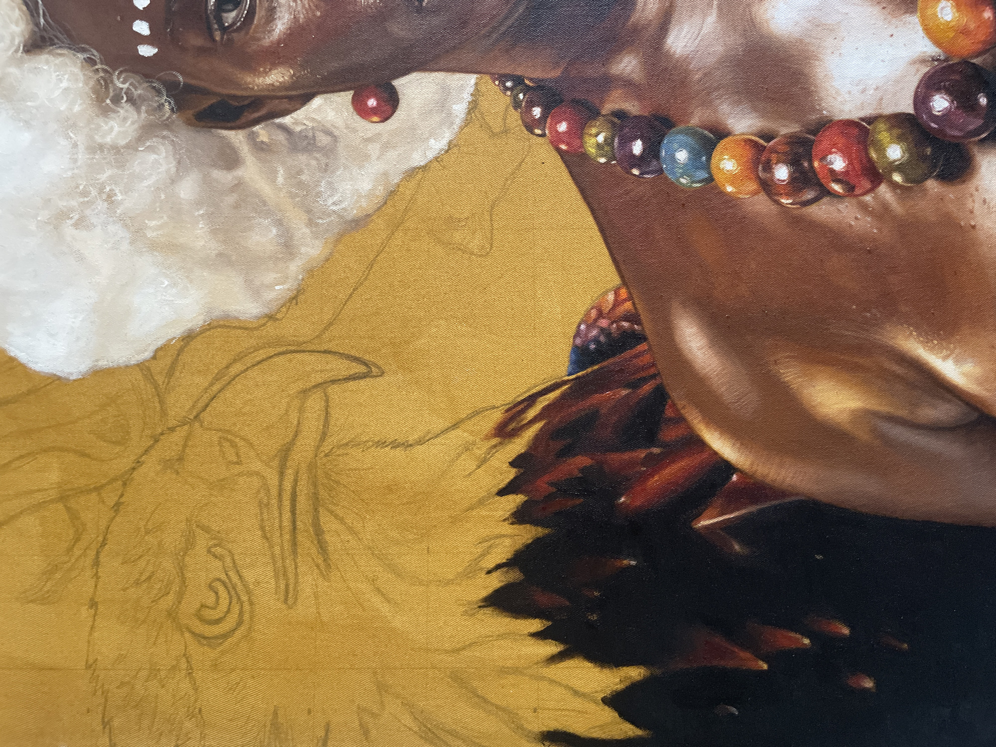This image depicts a portion of an oil painting on what appears to be a canvas or wooden panel. A dark-skinned person with white markings on their forehead and wearing a white, fluffy headpiece is partially visible at the top of the frame. They have a red earring and a multicolored beaded necklace around their neck.
The figure's skin is painted with a high level of detail, showing highlights and shadows. Beneath the necklace are dark red and black shapes which appear to be part of clothing or feathers.
The background is a solid golden color with faint pencil outlines of foliage or other elements not yet painted in. It is not possible to discern the location in Ado, Nigeria, from the image. There are no visible interactions or actions taking place. The image is focused on the partially finished portrait within a larger composition.
