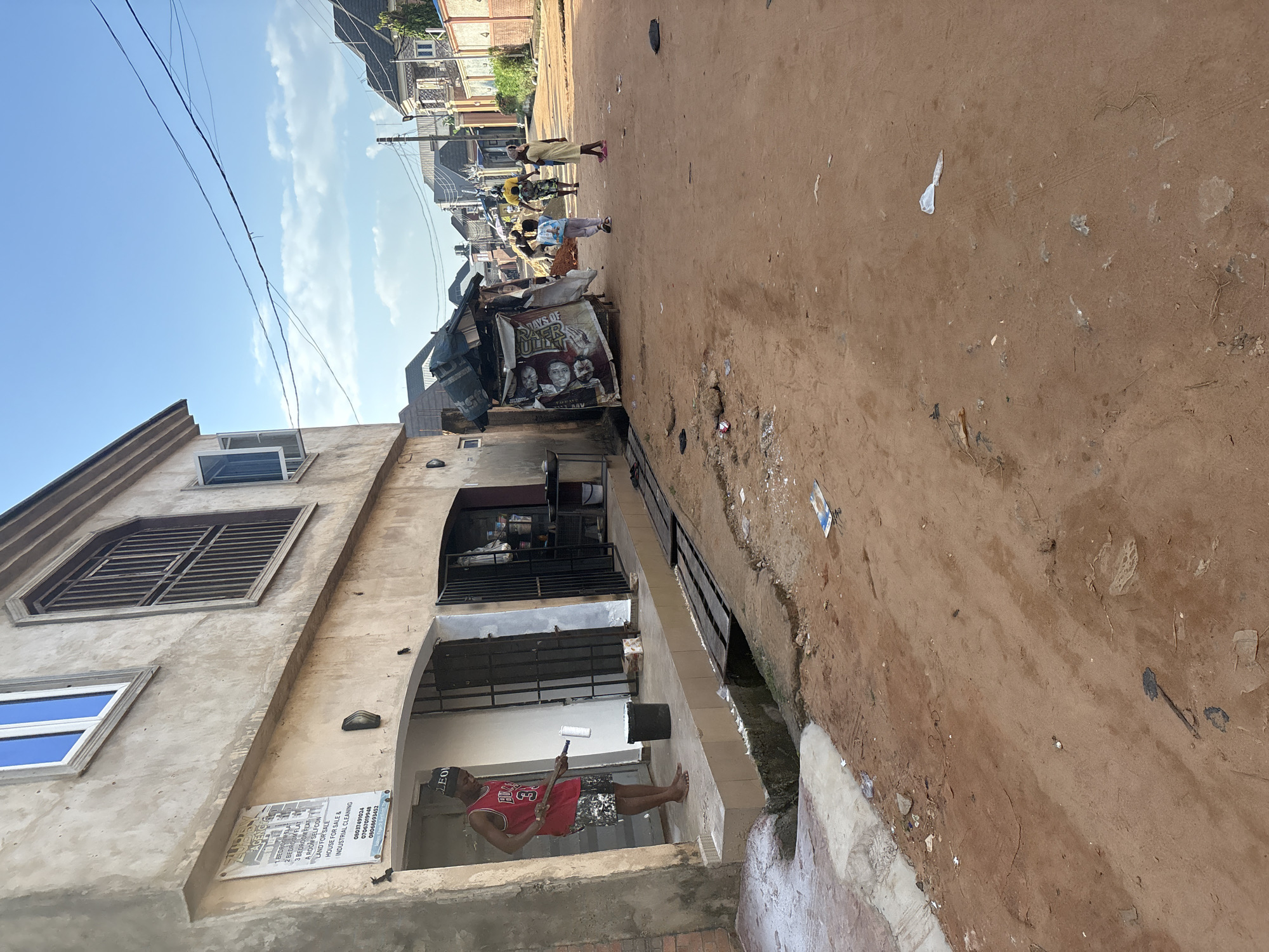 This image depicts a street scene in Obosi, Nigeria, under a bright, clear sky, suggesting it's daytime. The foreground is dominated by a dusty, reddish-brown dirt road, littered with small pieces of debris, indicating a somewhat unkempt environment. To the left, a multi-story building with a light-colored, textured facade is partially visible. It features several windows, one with blue glass and another with a prominent metal grate. A sign attached to the building's wall displays text related to property rentals and sales, including "1 BEDROOM FLAT," "2 BEDROOM FLAT," "3 BEDROOM FLAT," "LAND FOR SALE," and "HOUSE FOR SALE."
Further down the street, several people are scattered across the scene, some walking and others gathered near a makeshift stall or shop. This stall has a large banner advertising "DAYS OF SUPER BULL." The architecture of the surrounding buildings suggests a residential or mixed-use area. In the foreground, on a lower level of the building to the left, a man wearing a red basketball jersey and patterned shorts is painting a wall with a white roller. He appears barefoot. The overall atmosphere seems to be one of everyday life in a community, with elements of commerce and domestic activities visible.