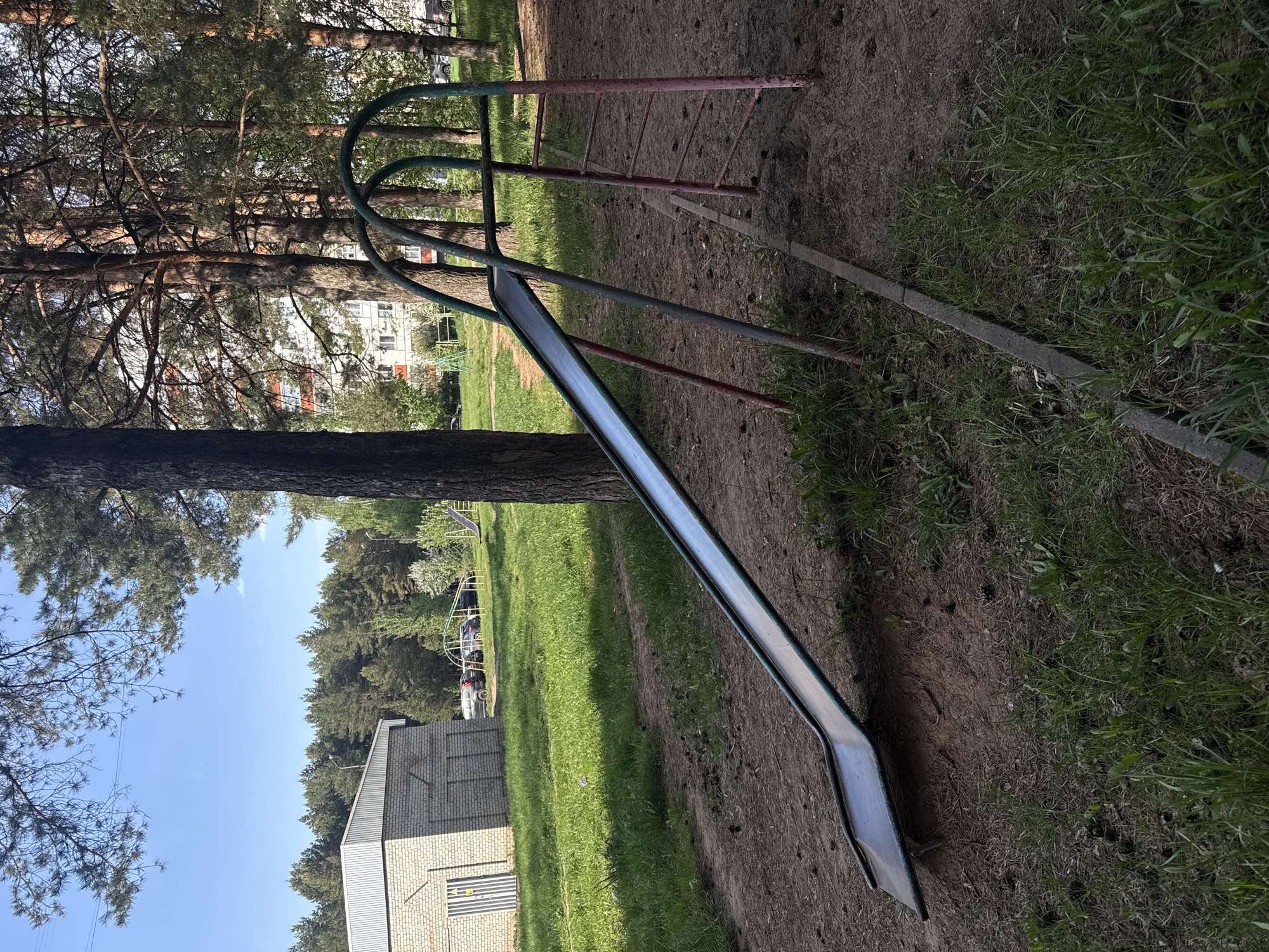 The image depicts a metal playground slide positioned adjacent to a tree, with its ladder section visible to the left of the frame. The slide sits on a patch of ground with interspersed grass and dirt. In the background is a building with a brick exterior. Trees are visible in the background, along with a residential building with multiple windows. The sky is a light blue with minimal clouds. The general location is Chaikovsky, Russia.