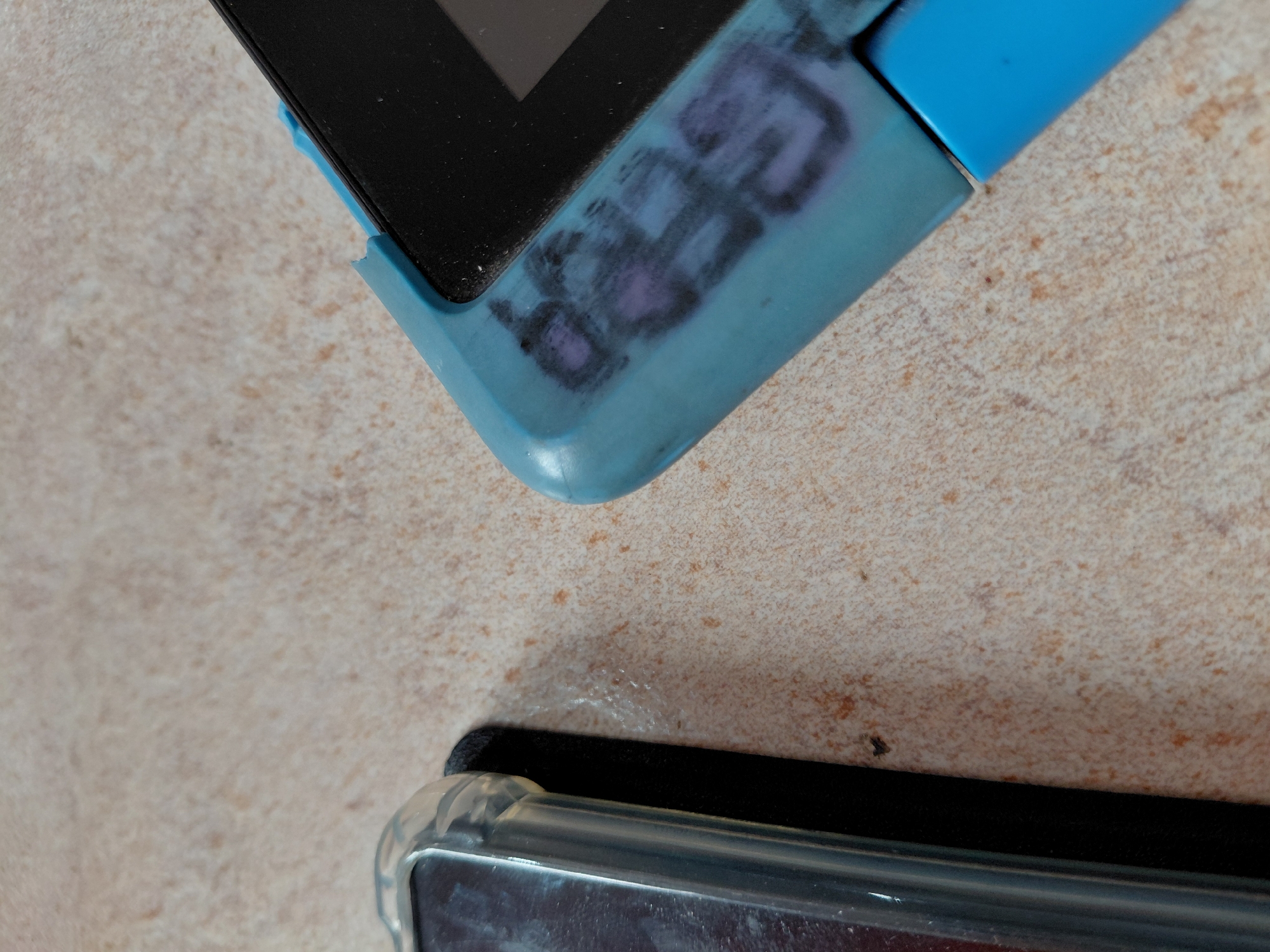The image displays a close-up view of two electronic devices resting on a light-colored, textured surface.
**Subjects:**
The primary subjects are two mobile electronic devices, likely smartphones or small tablets. The top device is partially visible, encased in a worn light blue protective bumper or case. Part of its black screen is also visible. Below it, at the bottom of the frame, another electronic device is partially visible, encased in a clear, transparent protective case. The background is a light beige or cream-colored surface with brown speckles, possibly a countertop or tiled floor.
**Activity or event taking place:**
No activity or event is taking place; the devices appear to be stationary, simply resting on the surface.
**Notable details:**
The time of day is not discernible, but the lighting suggests an indoor setting. The surface on which the devices rest has a speckled, granular appearance, resembling light-colored ceramic tiles or a laminate countertop. The blue case of the upper device shows significant wear and tear, including scratches and scuff marks. One corner of the blue case appears to be chipped or damaged. The screen of the device in the clear case at the bottom appears somewhat dusty or smudged.
**Visible text:**
On the light blue casing of the upper device, there are distinct, faded markings in black and purple. These marks appear to spell out "SBEG" in a somewhat stylized, graffiti-like font, though the letters are not perfectly clear due to fading and the texture of the case.
