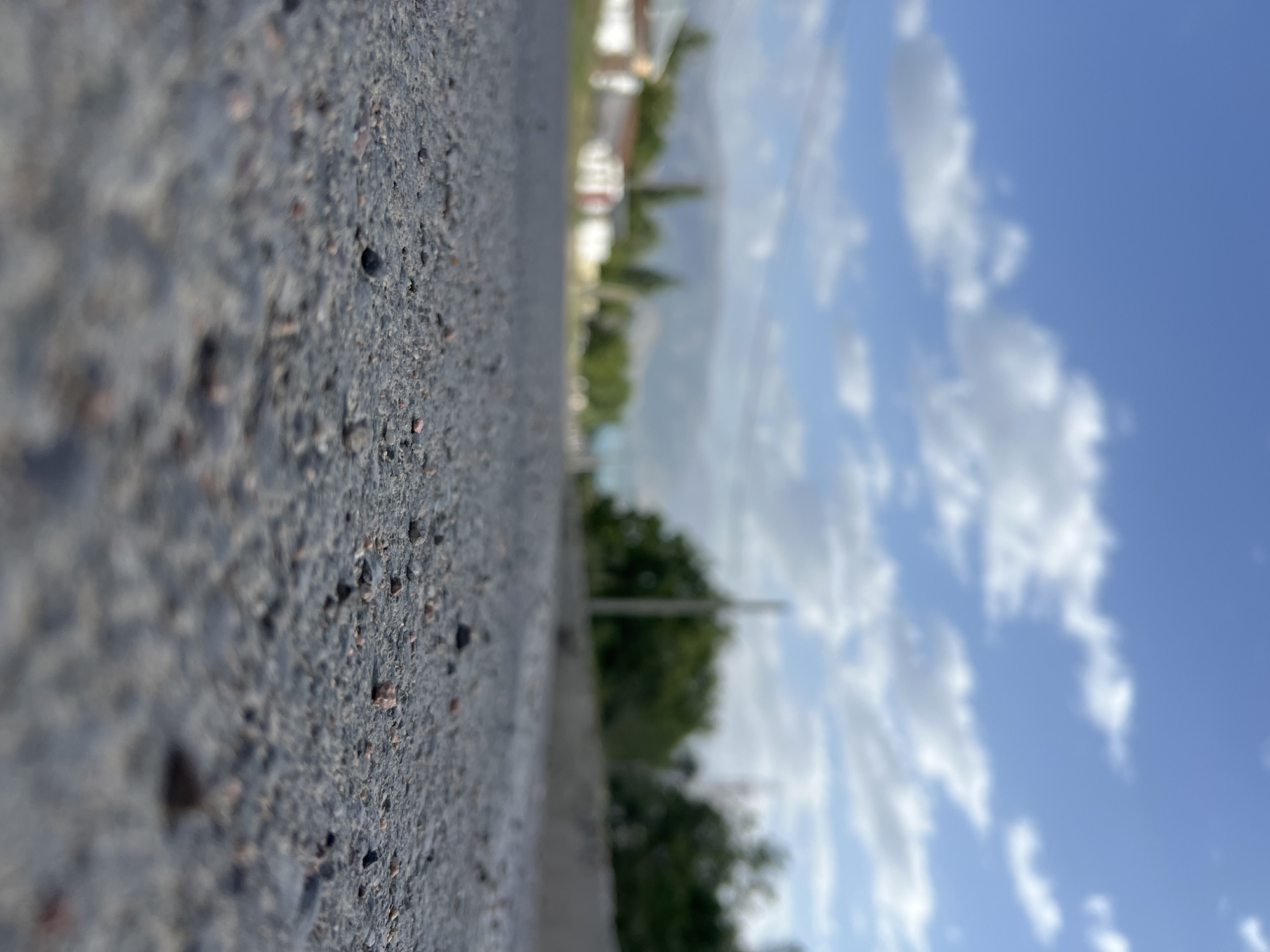 An extremely low-angle, close-up shot focuses on the textured surface of an asphalt road in Yusuphana, Uzbekistan. The asphalt is composed of small, dark gray stones with occasional flecks of lighter gray and reddish-brown. In the foreground, the road surface is sharp and detailed, showing individual pebbles. As the eye moves towards the background, the scene becomes progressively out of focus.
Beyond the road, a blurred landscape unfolds under a bright blue sky dotted with fluffy white clouds. A low, out-of-focus line of buildings, likely houses, runs along the left side of the road. Some of these buildings appear to have white walls and red or white roofs. Trees with lush green foliage are visible behind the buildings and along the right side of the road, forming a soft, blurred mass. In the far distance, a hazy mountain range can be discerned. A utility pole is visible as a faint vertical line among the trees. The overall impression is a serene, daytime scene in a semi-urban or rural environment. There are no people or animals visible in the image.