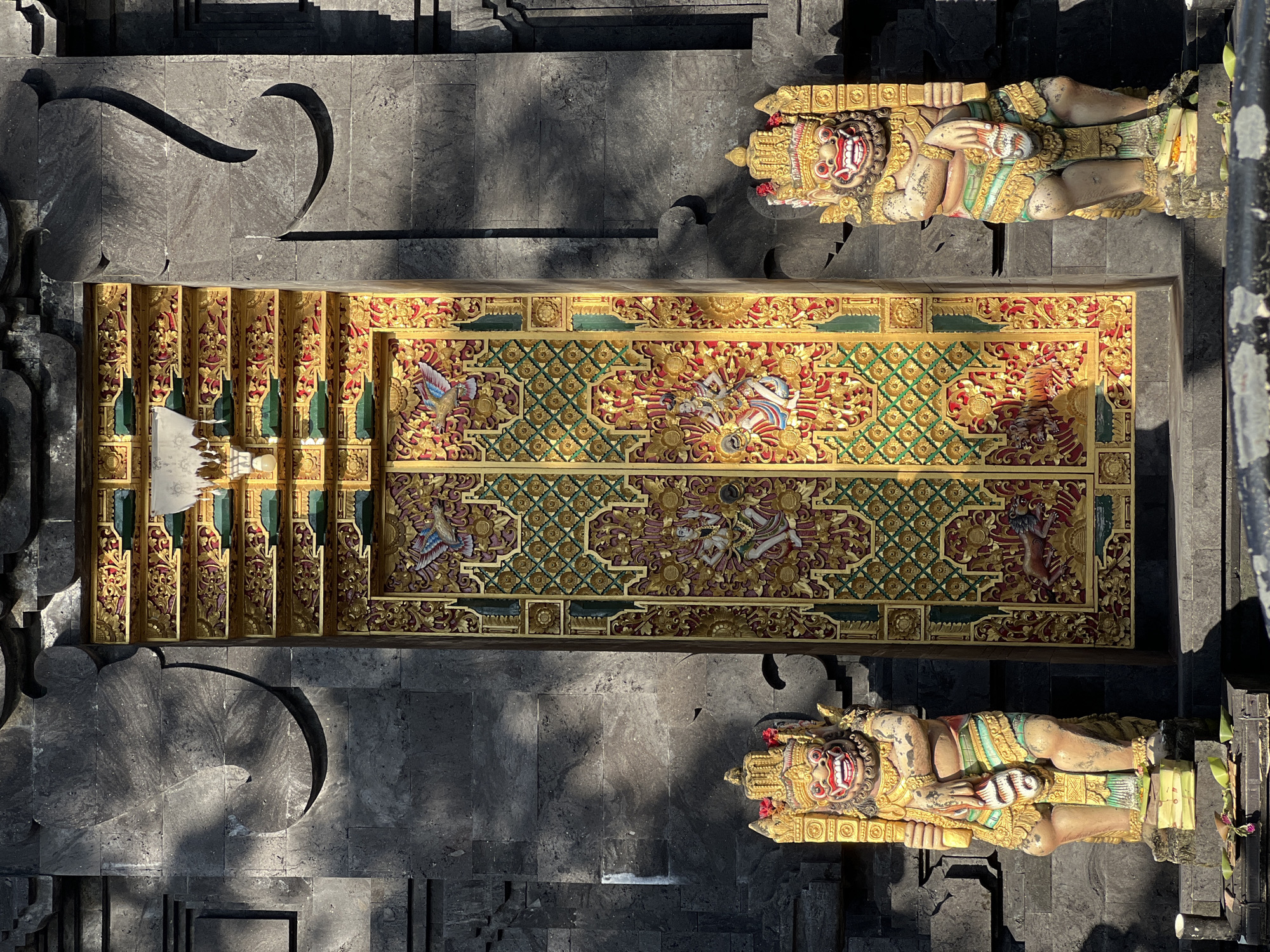 An overhead shot captures a richly adorned entrance or ceremonial pathway, likely in a temple complex in Kuta, Indonesia. The central feature is a highly elaborate, gilded wooden structure, possibly a gate, door, or section of stairs, featuring intricate carvings. This central piece is dominated by gold, red, and green hues, depicting stylized mythological figures, birds, and geometric patterns. Two large, ornate guardian statues, characteristic of Balinese art (likely Dwarapala figures), flank the central structure. These statues are depicted lying horizontally, with fierce, red-painted faces, prominent fangs, and elaborate golden headdresses. Their bodies are adorned with colorful garments in shades of gold, green, and red, and they appear to be holding staffs or clubs.
The entire scene rests on a dark grey, textured stone ground, possibly volcanic rock, which extends beyond the central area. Strong, defined shadows are cast across the stone, including large, abstract curved shapes, indicating bright, direct sunlight. The lighting suggests it is midday or early afternoon under a clear sky. No discernible human activity is taking place, presenting a static view of the architectural and sculptural elements. There is no visible text in the image.