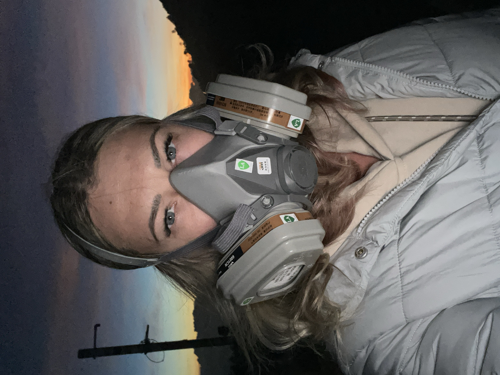 A woman with blonde hair wearing a grey respirator mask and a puffy jacket is facing the camera. Behind her, the sky transitions from dark blue to orange, resembling a sunset or sunrise. A utility pole is partially visible in the lower left corner. The location is unknown.