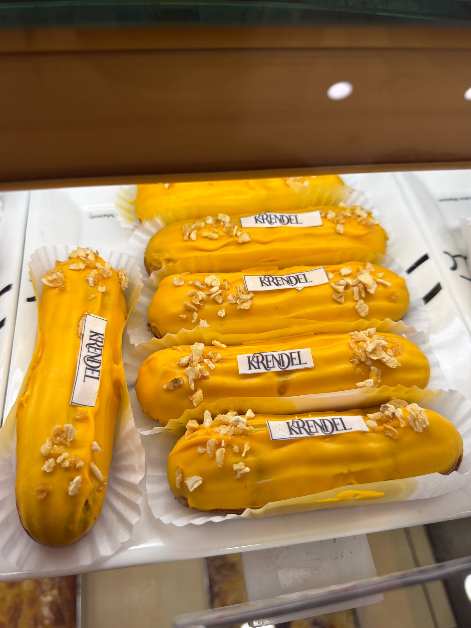 Six yellow eclairs are arranged on a white tray. Each eclair is topped with small, light-brown crunchy pieces and has a white paper label with the word "KRENDEL" printed in black. The eclairs are positioned diagonally from the bottom left to the top right of the image. The background appears to be a display case with a wooden frame at the top. The image was taken in Pavlodar, Kazakhstan.