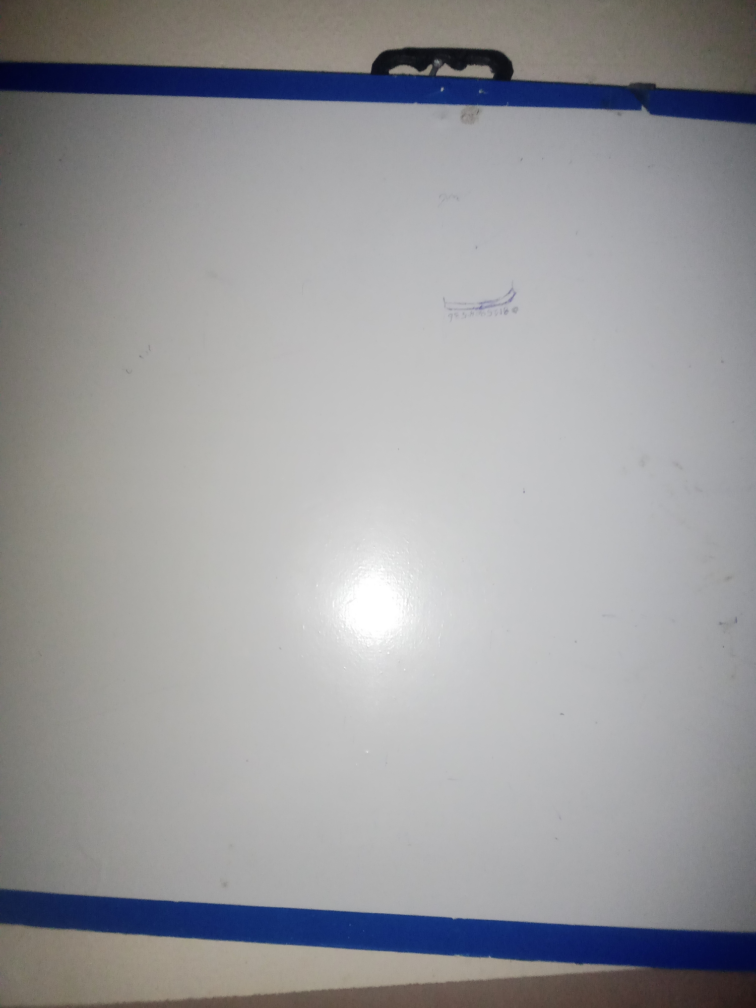 The image shows a whiteboard with a blue border, resting on a surface that appears to be a table or desk. There is a black handle attached to the top of the whiteboard. Faint markings are visible on the whiteboard's surface, including what looks like a curved line with some numbers underneath it. The lighting suggests an indoor environment, with a bright reflection on the whiteboard's surface indicating a light source. The overall scene is static, with no active subjects or interactions depicted. The location in Jauro Yinu, Nigeria, is an external context not visible within the image itself.
