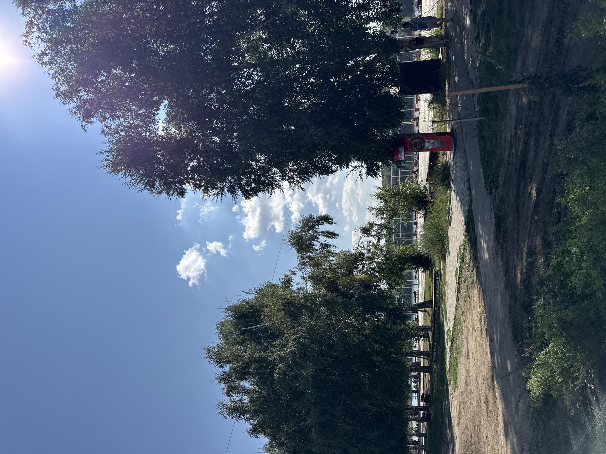 The image depicts an outdoor scene, likely a park or recreational area, with lush green trees dominating the foreground and midground. The sky is a clear blue with scattered white clouds, and the sun is shining brightly, casting shadows. In the midground, there's a paved walkway and a dirt path, with several benches visible. Further back, a body of water, possibly a river or lake, can be seen. People are scattered throughout the area, some sitting on benches, others walking. A distinctive red structure resembling a vintage phone booth stands near the center of the image. The background shows more trees and a structure that appears to be a building or a pavilion. The overall atmosphere is one of a relaxed, sunny day in a public outdoor space. The specific location in the city of Oskemen, Kazakhstan, is not explicitly identifiable from the visual cues in the image alone.
