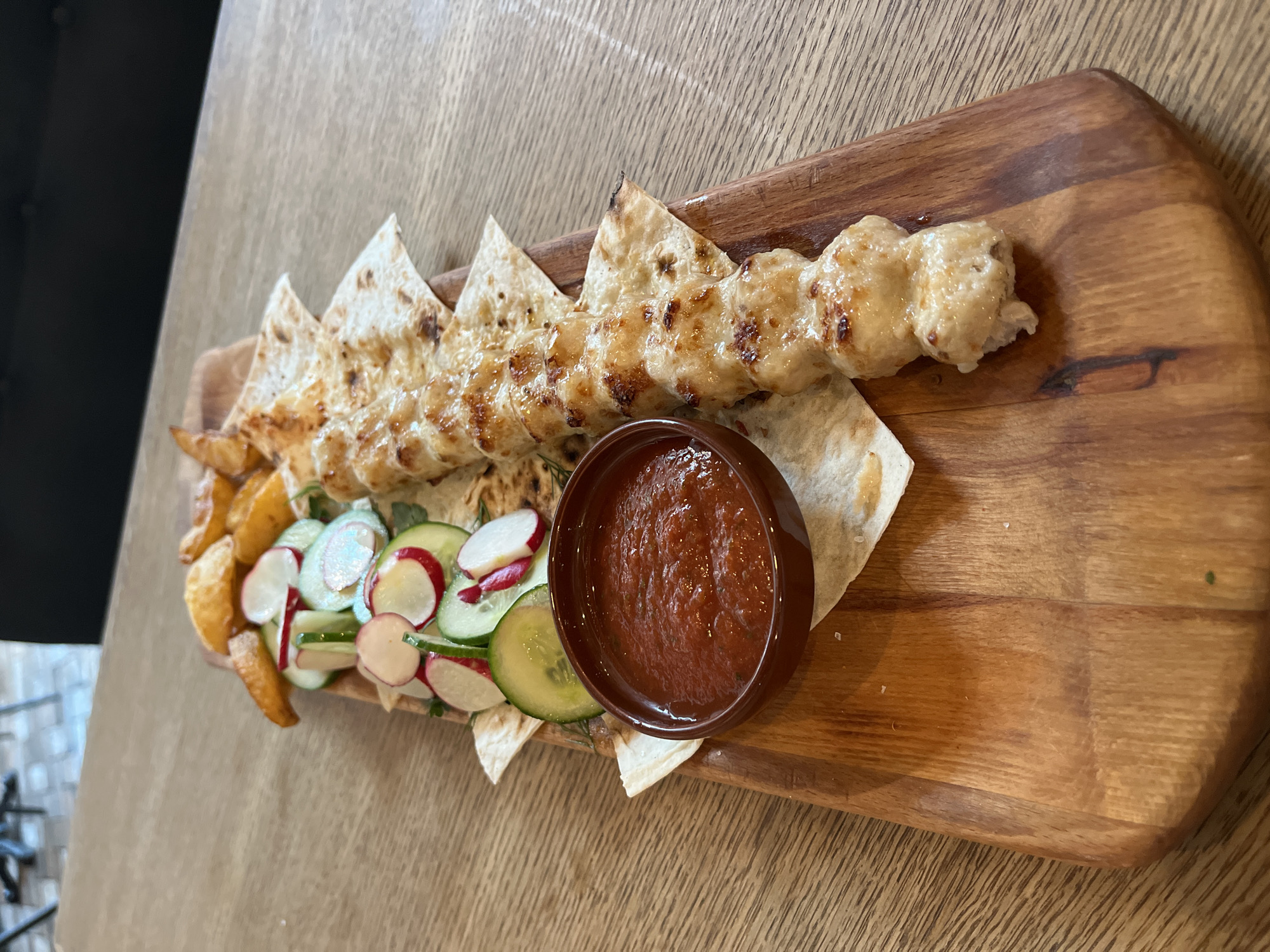 The image presents an appetizing meal artfully arranged on a rustic wooden serving board, likely in a restaurant or cafe in Sochi, Russia.
The central dish features a long piece of skewered meat, possibly chicken or fish kebab, characterized by its light color, glossy glaze, and faint char marks, indicating it has been grilled or baked. This main component rests upon several triangular pieces of flatbread, resembling lavash, which also exhibit light grilling.
To the left of the main dish, a side of golden-brown potato wedges is visible. A fresh and colorful salad accompanies the meal, consisting of thinly sliced green cucumbers and bright red radishes. A small, dark brown ceramic bowl is placed near the center, holding a rich red sauce, likely a tomato-based condiment or adjika, with a slightly textured consistency.
The wooden serving board features a visible grain and rounded edges, contributing to the rustic aesthetic of the presentation. The background consists of a blurred wooden table surface, with a dark, indistinct area to the far left. The lighting is bright and even, suggesting an indoor setting during daylight hours. No people are visible, and there is no discernible text in the image. The arrangement implies a meal ready for consumption, emphasizing its visual appeal and components.