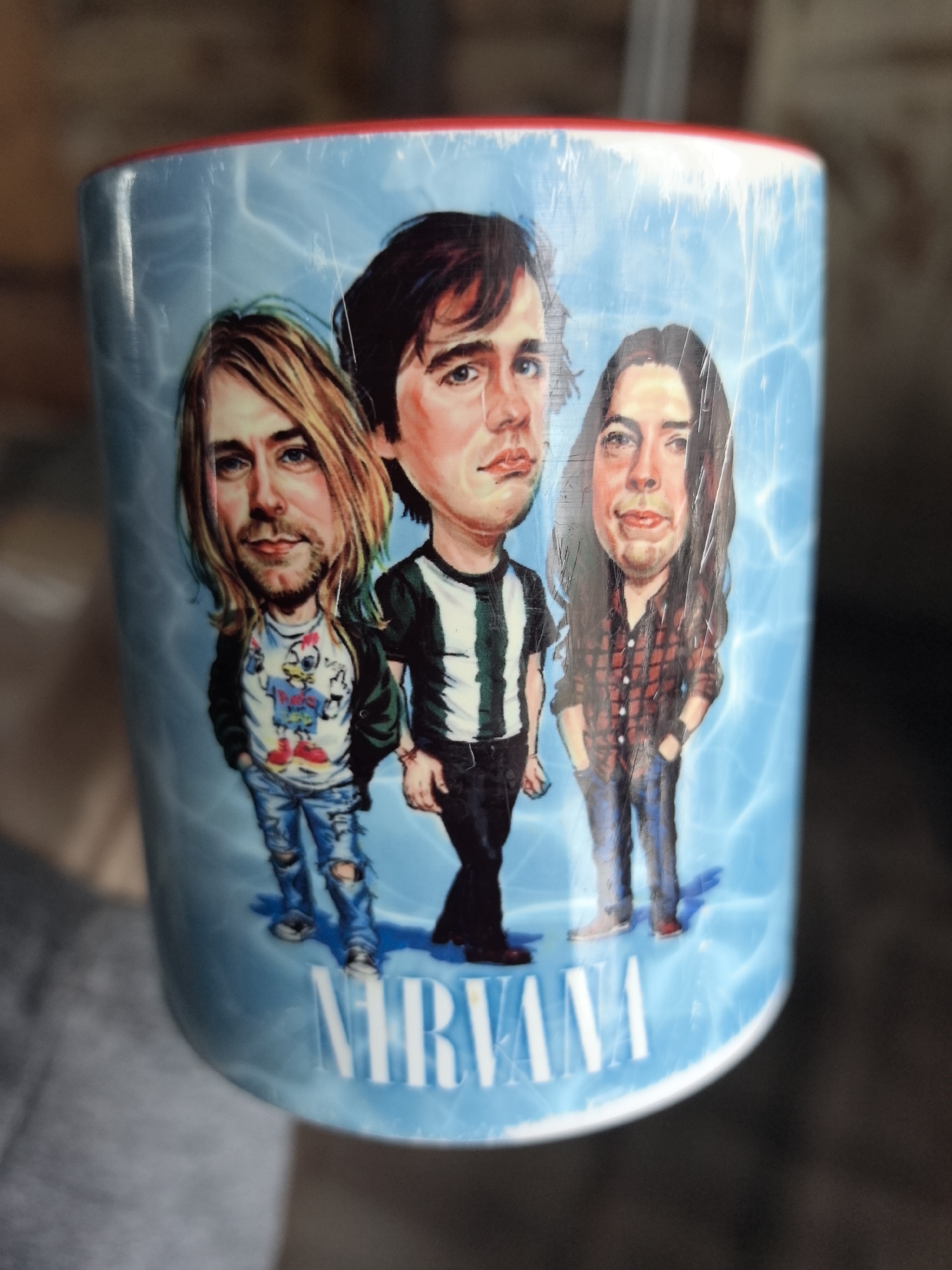 A ceramic mug featuring caricatures of the band Nirvana is depicted, held by an unidentifiable individual. The mug's exterior is light blue with a subtle wavy pattern, displaying three cartoonish figures: Kurt Cobain (left, blonde hair, beard, wearing a chicken-design t-shirt, dark jacket, and ripped jeans), Krist Novoselic (center, dark hair, clean-shaven, wearing a striped shirt and dark pants), and Dave Grohl (right, long dark hair, beard, wearing a plaid flannel shirt and jeans). The word "NIRVANA" in white capital letters is printed below the figures. The mug's top rim is red. Numerous fine scratches are visible across the printed surface of the mug, particularly over the figures. A blurred human hand, wearing a grey sleeve, holds the mug on the left side. The background is dark and heavily out of focus, providing no discernible details of the immediate environment. The precise location within the city of Noviy, Russia, cannot be determined from the visual information present in the image.