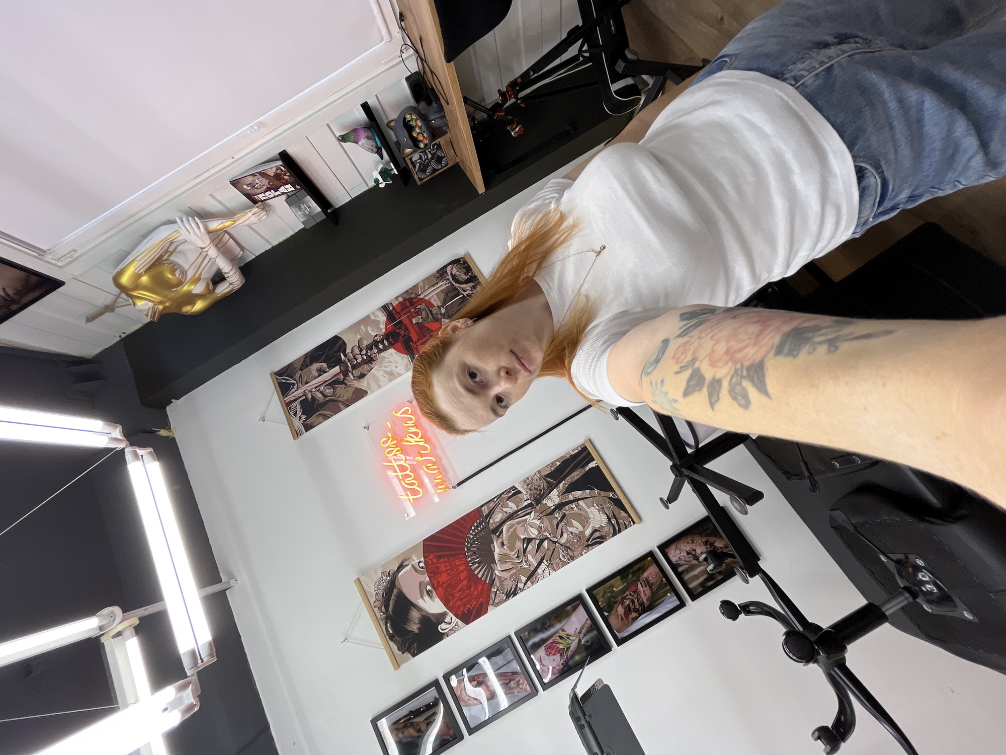 The image depicts a selfie taken by a woman with long, red hair inside what appears to be a tattoo studio. She is wearing a white t-shirt and blue jeans. Her left arm, visible in the foreground, has tattoos. The background shows a white wall with various framed art pieces and scrolls, one featuring a woman with a red fan. A neon sign in orange reads "tattoos_markus". There is also a mannequin torso with gold paint, a desk with office supplies, and a black exercise bike. The lighting is bright, with fluorescent tubes visible on the ceiling. The scene is located in a studio.