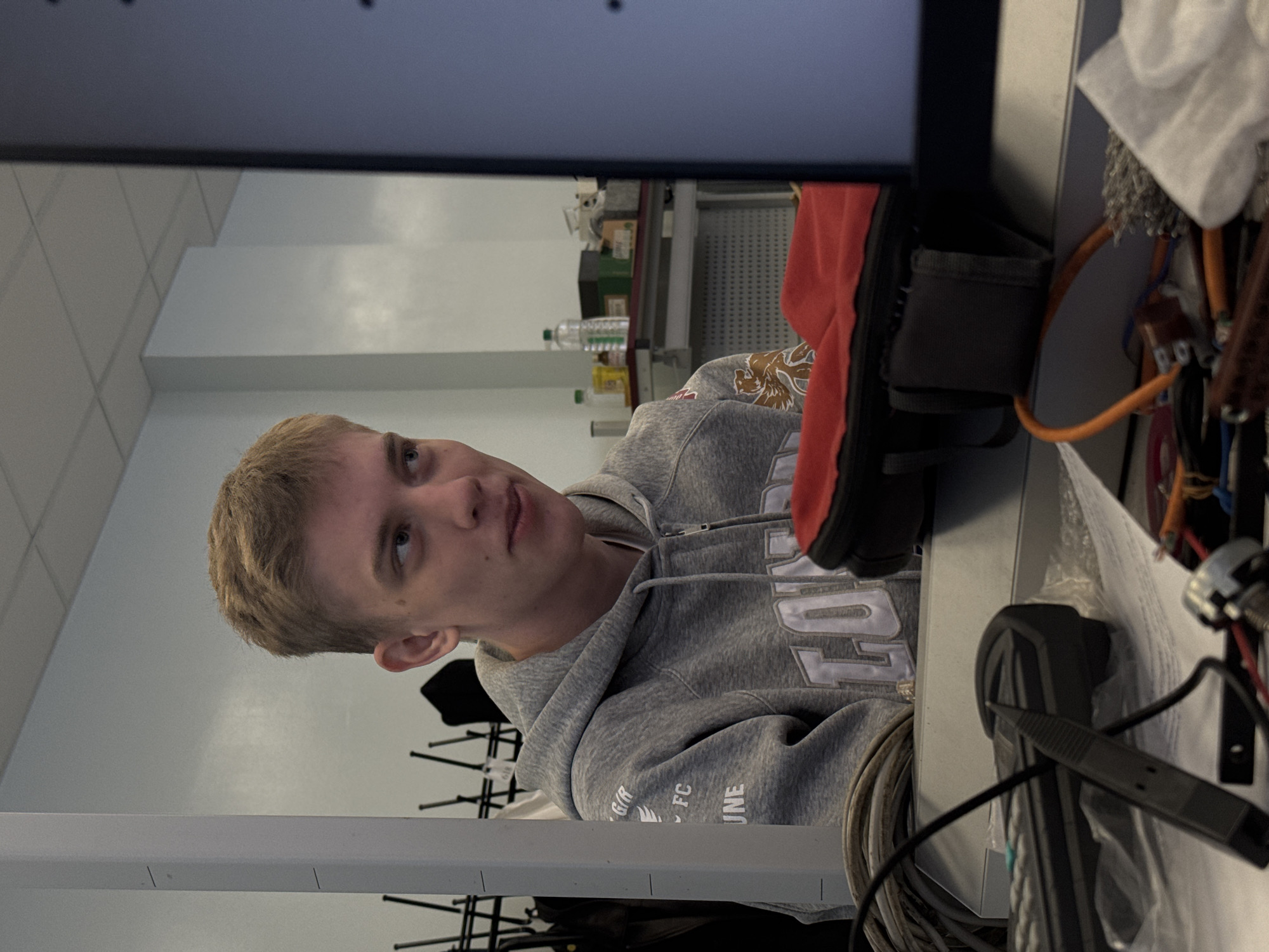 A young man with short blonde hair and wearing a grey hoodie sits at a desk, partially obscured by a computer monitor. On the desk are various items including a red shoe, wires, tools, and papers. In the background are white walls with visible electrical conduits and a shelving unit containing bottles and containers. There is no visual information to confirm or deny the location in the city of Perm, Russia.