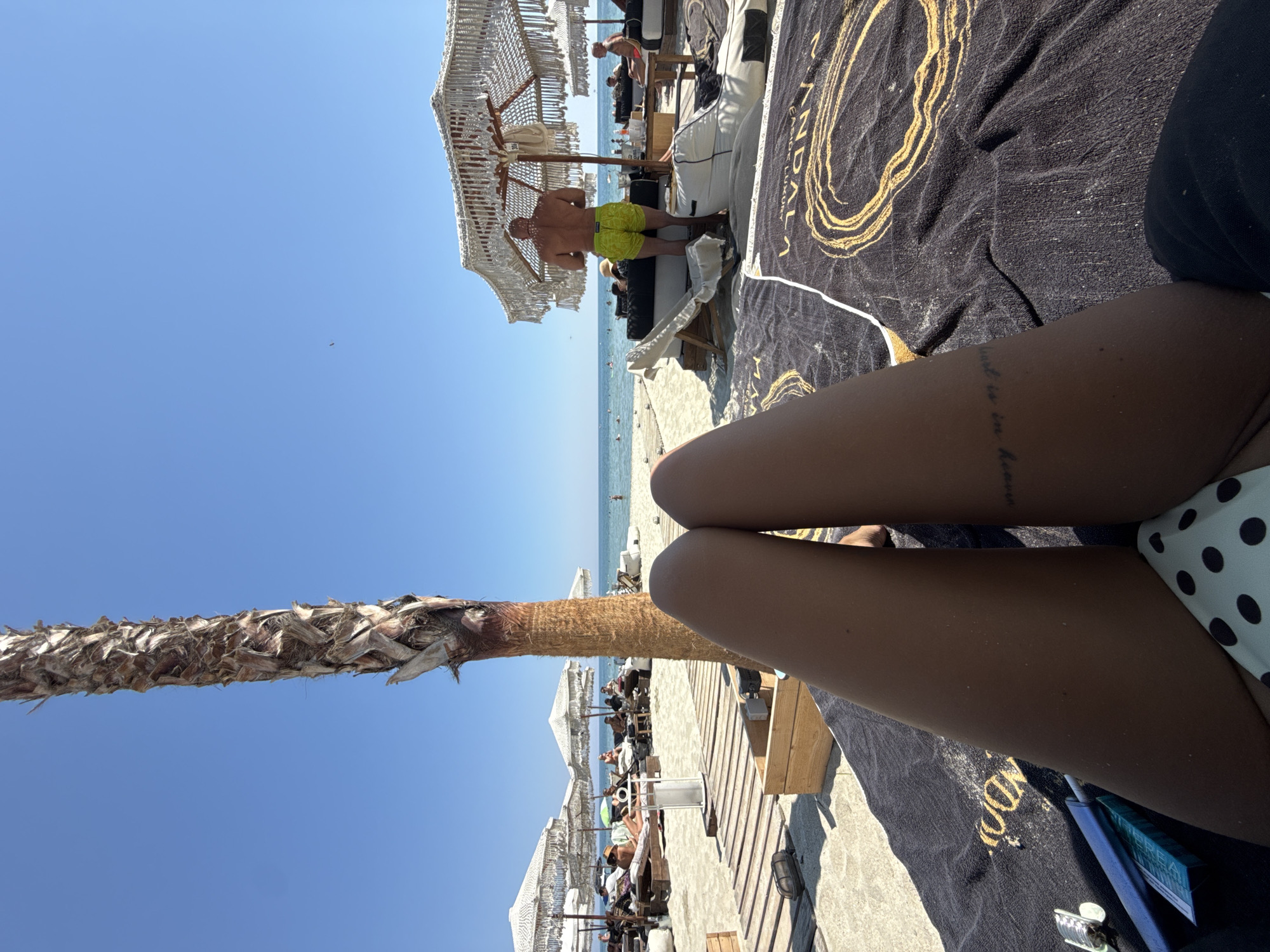 This is a first-person perspective, eye-level, medium shot of a person's legs and bikini bottom on a beach towel on a sunny day. The person is lying on a black towel with a gold circular design. To the left, a blue package and a metallic object are visible. The person's legs are tanned and show a subtle tattoo on the inner thigh. In the background, a wooden walkway leads to a beach with numerous lounge chairs and umbrellas. A palm tree is prominently featured in the foreground, to the left of the person's legs. The sea is visible in the distance, with a few people swimming. The sky is clear and blue. The scene appears to be at a beach resort in the city of Katerini Municipality, Greece, likely on a summer day given the bright sun and beach setting.