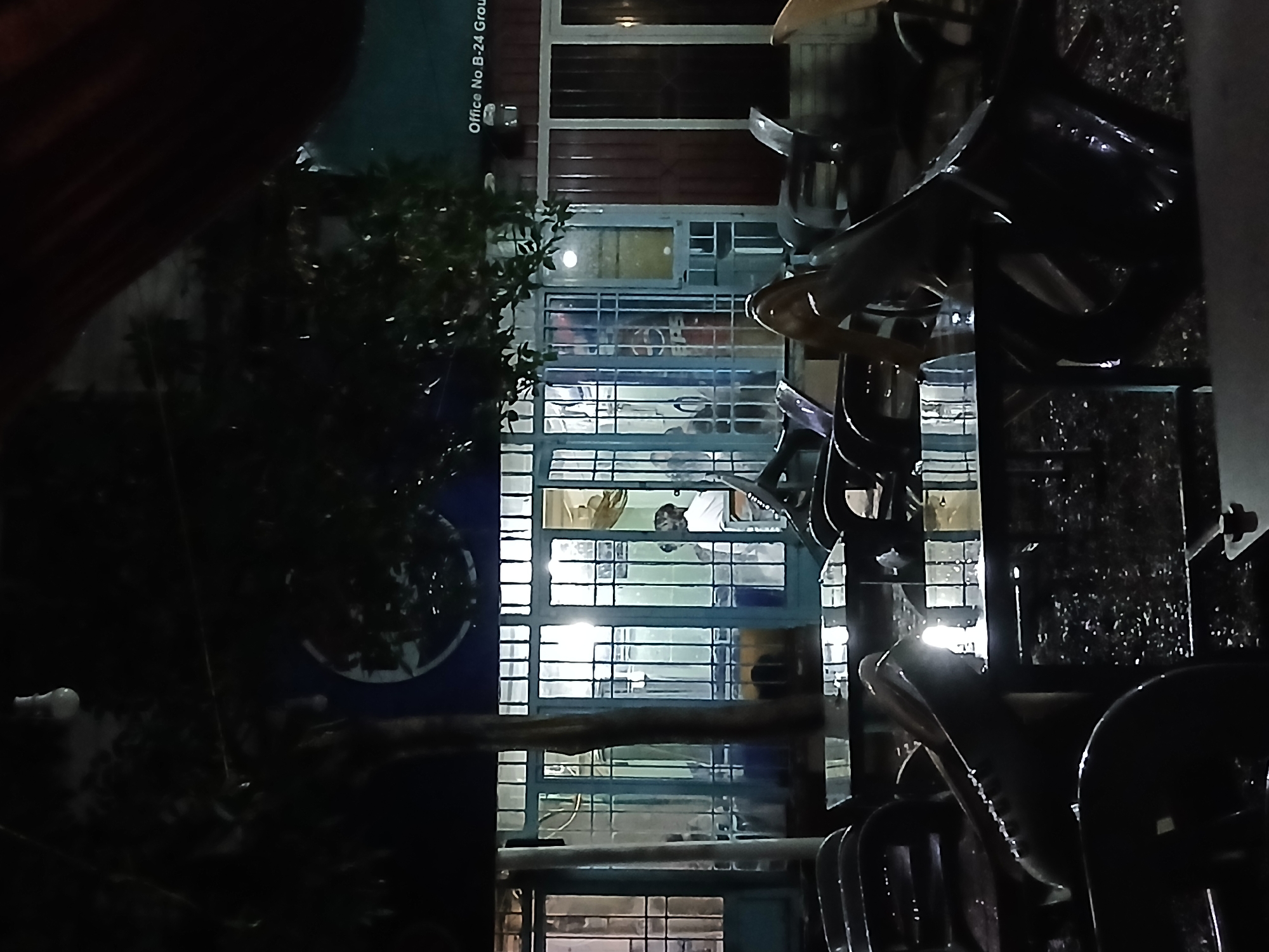 The image depicts the exterior of what appears to be an office building at night in Quetta City Tehsil, Pakistan. A sign on the building reads "Office No B-24 Group". The building has a grid-like window facade behind a security grill. Inside, figures can be seen in what looks like an office environment with overhead lighting. The foreground includes objects that appear to be chairs or seating, possibly stacked, with reflective, wet surfaces, suggesting it may have rained or there is moisture present. To the left of the building, there is foliage, potentially a tree or shrub, with a sign with a logo. The overall lighting is low.