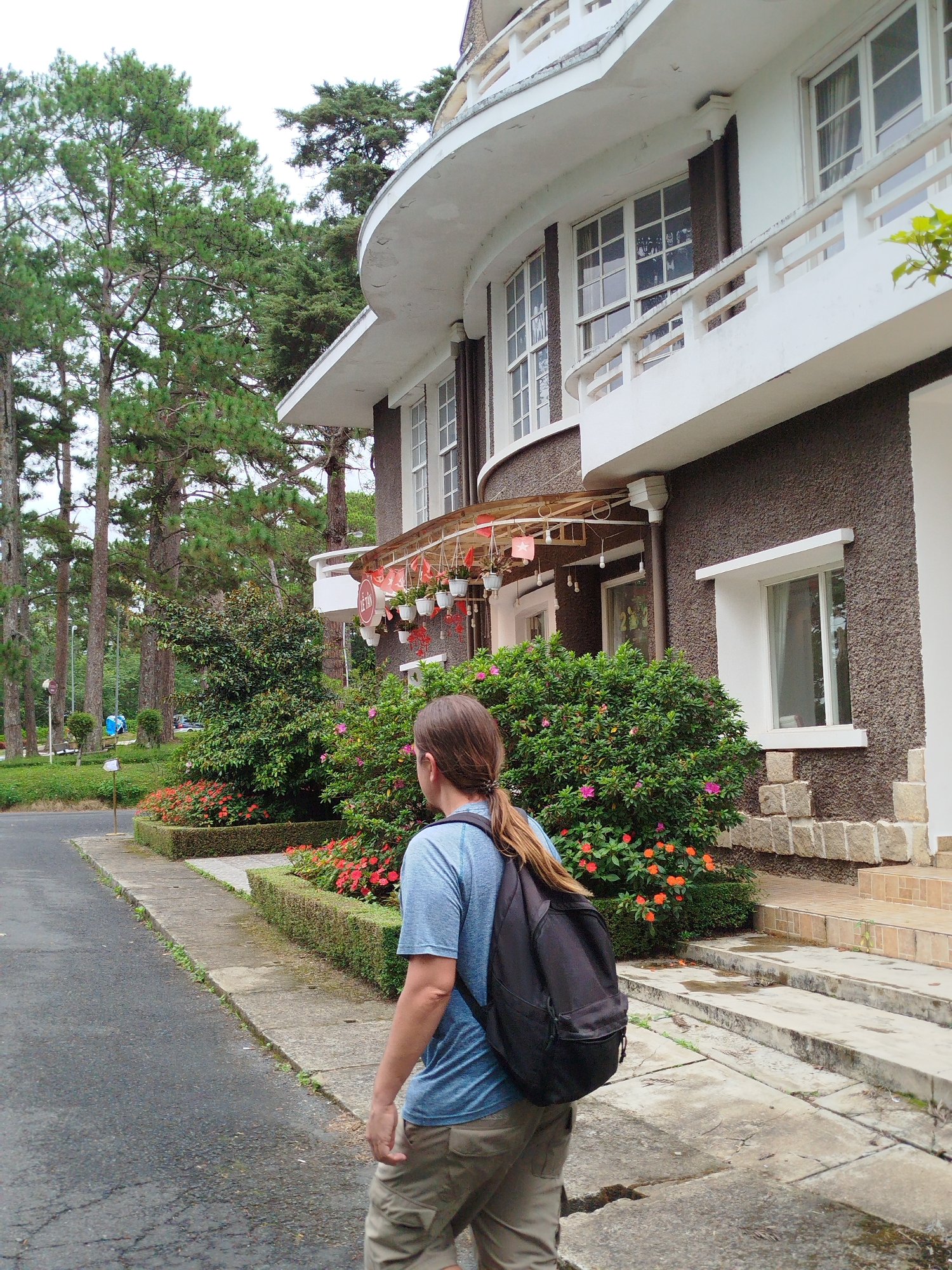 In Da Lat, Vietnam, a person with long brown hair, wearing a blue t-shirt and khaki shorts, walks along a paved path next to a dark brown, textured building with white trim and multiple windows. The building has a distinctive curved architectural feature and a balcony. The path is lined with manicured hedges and a vibrant display of red and orange flowers. Tall pine trees are visible in the background, creating a natural, verdant setting. A black backpack is worn by the person. The ground appears wet in places, suggesting recent rain.