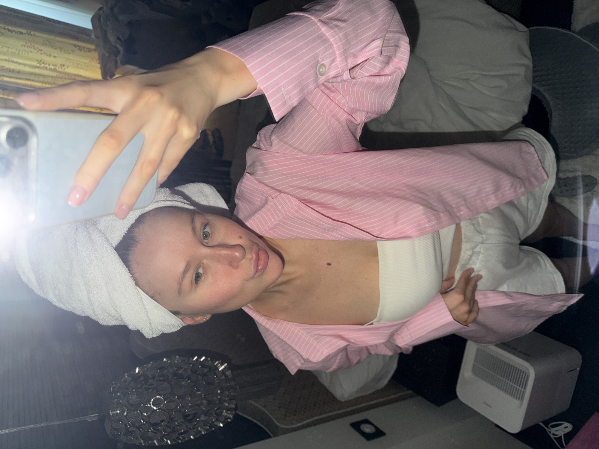 A young woman is taking a selfie in what appears to be a domestic setting. She has a white towel wrapped around her head and is wearing a white tube top with a pink and white striped button-up shirt open over it. She is holding a smartphone up to take the photo.
Around her, there is a bed with pillows, and a small white air purifier is visible on the floor. A decorative metallic object, possibly a lamp or bowl, is on a table or surface below her.
There's no information in the image to suggest this location is in the city of Khimki, Russia.