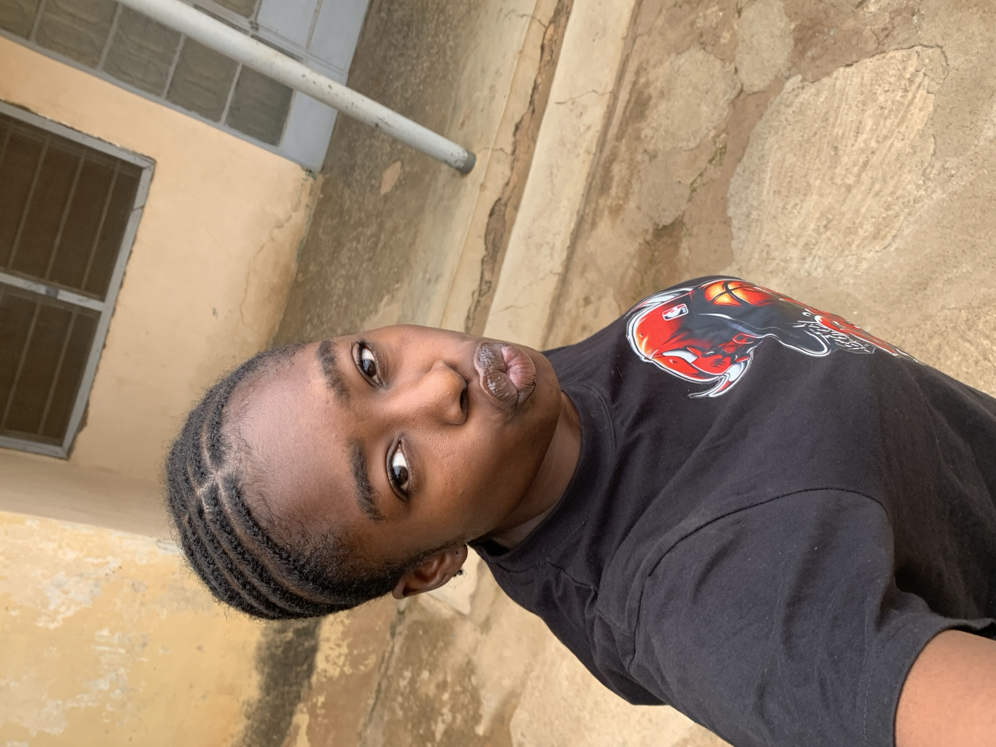 A person with dark skin and braided hair is facing the camera with puckered lips. They are wearing a black t-shirt with a red graphic design on the shoulder area. The background appears to be an outdoor location with light-colored concrete and a building with barred windows. The specific location is Rafin Par, Nigeria.