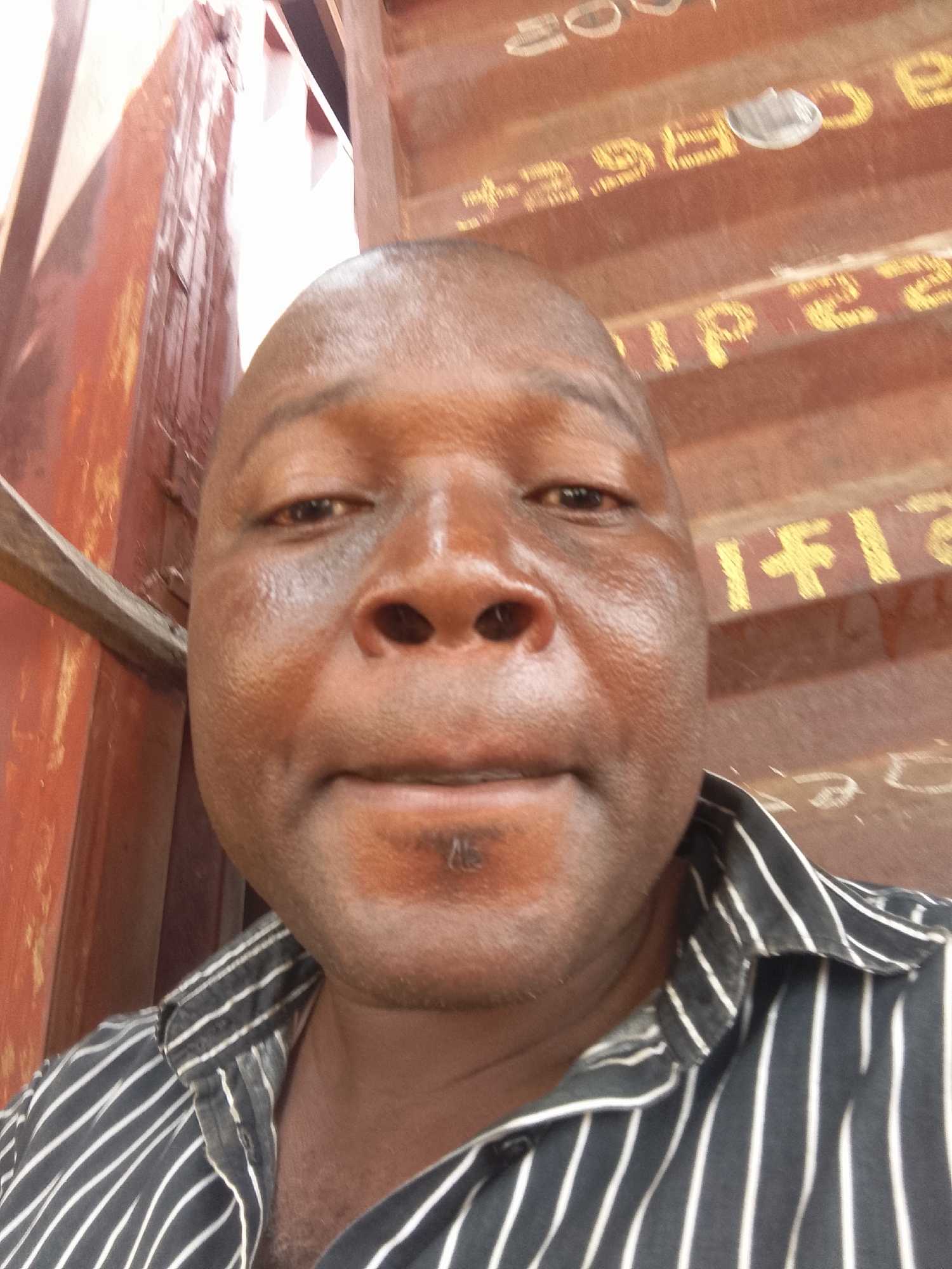 A close-up selfie of a Black man with a shaved head is positioned in front of a blurred background of a cargo container. The man is wearing a black and white striped shirt. The cargo container is predominantly reddish-brown with yellow lettering. The location is presumed to be the city of Unknown, Unknown.