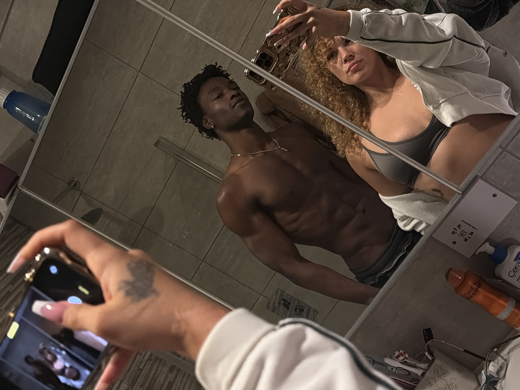 The image depicts a mirror selfie taken by a woman in a bathroom, presumed to be in London, United Kingdom. The reflection shows a light-skinned female in the foreground, with curly light brown hair, wearing a grey sports bra and an open light-colored zip-up jacket. Her right hand, featuring a French manicure and a dark tattoo on its back, holds a gold-cased smartphone used to capture the image. A dark-skinned male is reflected standing behind the woman. He has short dark curly hair, a muscular shirtless physique, and wears dark shorts along with a gold chain and pendant. Both individuals appear to be posing for the photograph. The bathroom environment features light-colored tiled walls and a partially visible glass shower enclosure. A counter below the mirror displays a pair of glasses, an orange L'Oréal bottle, a white and blue CeraVe pump bottle, and a container holding toothbrushes. A piece of paper with Cyrillic text is affixed to the wall next to these items. An upper shelf is reflected, showing a blue water bottle and a black bag. A brown towel is draped near the male's right shoulder in the reflection.