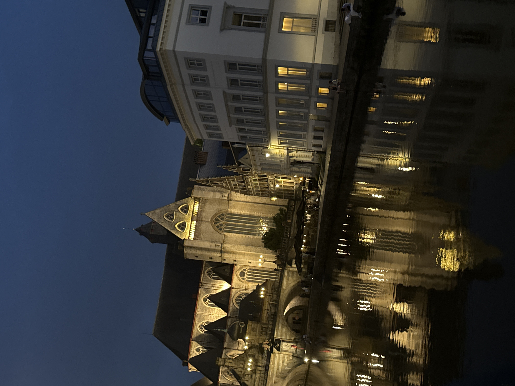 An evening shot shows a canal with reflections of illuminated buildings on its surface. On the left, a large Gothic cathedral, likely St. Bavo's Cathedral in Ghent, Belgium, is visible with its arched windows and pointed spires. Adjacent to it, on the right, is a more modern, white building with multiple rectangular windows. The sky is a deep blue, indicating it is twilight or nighttime. Several people are seated on the canal's edge, and others are dining at outdoor tables, suggesting a public space or promenade. The overall scene depicts a European city at night, characterized by historic architecture and a calm waterway.