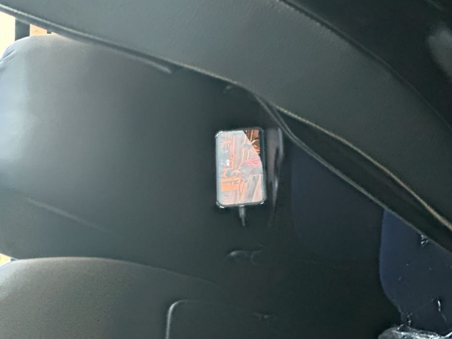 An eye-level shot reveals the interior of a vehicle, likely a car. A dark, padded headrest occupies the left side of the frame, while a portion of a dark dashboard or console stretches across the top. Adhered to the back of the headrest is a smartphone in a clear case, appearing to be charging via a cable. The phone screen displays a blurry image, possibly a video or live feed. A navy blue portion of a seat is visible in the bottom right.