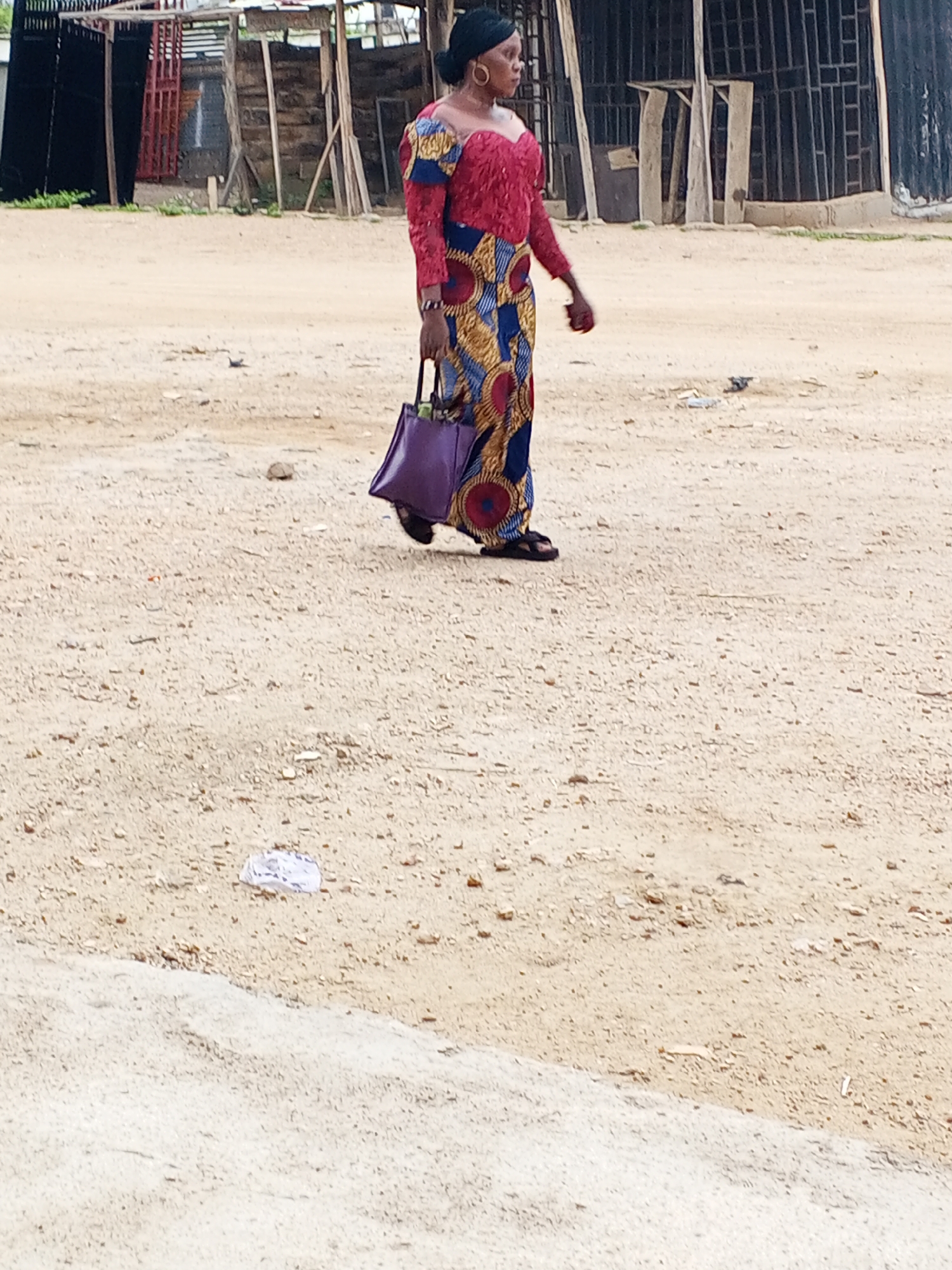 In Otokiti, Nigeria, an individual wearing a headwrap, a dress with a red lacy top and a blue and gold patterned skirt, and sandals, is walking across a dirt and sandy terrain. The individual is carrying a purple bag. The background shows wooden structures and fencing. Debris is scattered on the ground.