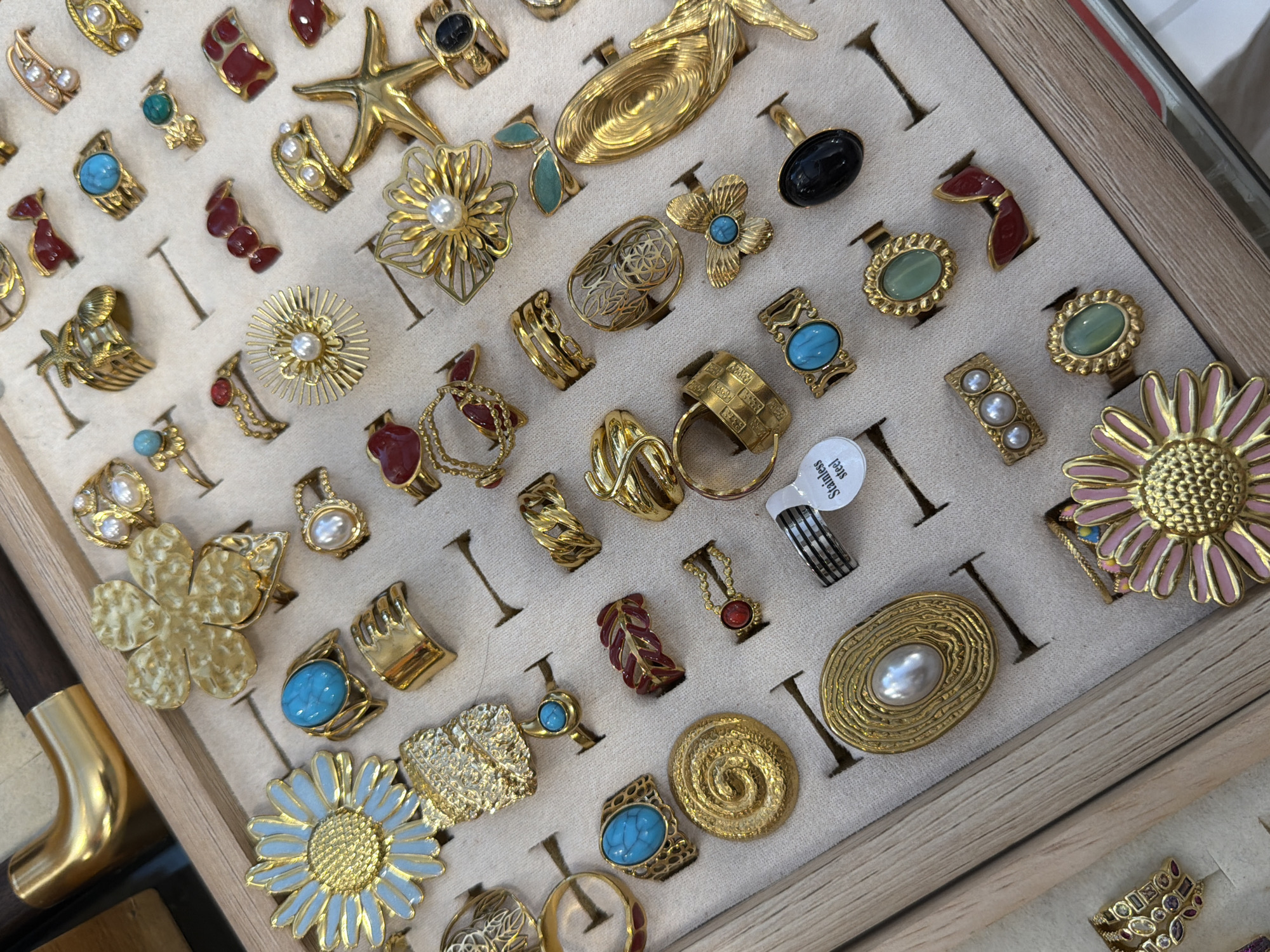 An overhead, slightly angled shot shows a jewelry display case filled with numerous rings. The rings are predominantly gold-toned and feature a variety of designs, including floral motifs, abstract shapes, and gemstone embellishments in colors like turquoise, red, black, and green. Some rings have pearls or small, faceted stones. The display case itself appears to be made of wood with a light-colored fabric lining, divided into compartments to hold each ring. In the upper left corner, a portion of a golden handle or decorative element with a dark wood inlay is visible, suggesting it might be part of a larger display or furniture. The overall scene is well-lit and focuses on the intricate details of the jewelry. The image is cropped to prominently feature the rings and their arrangement.