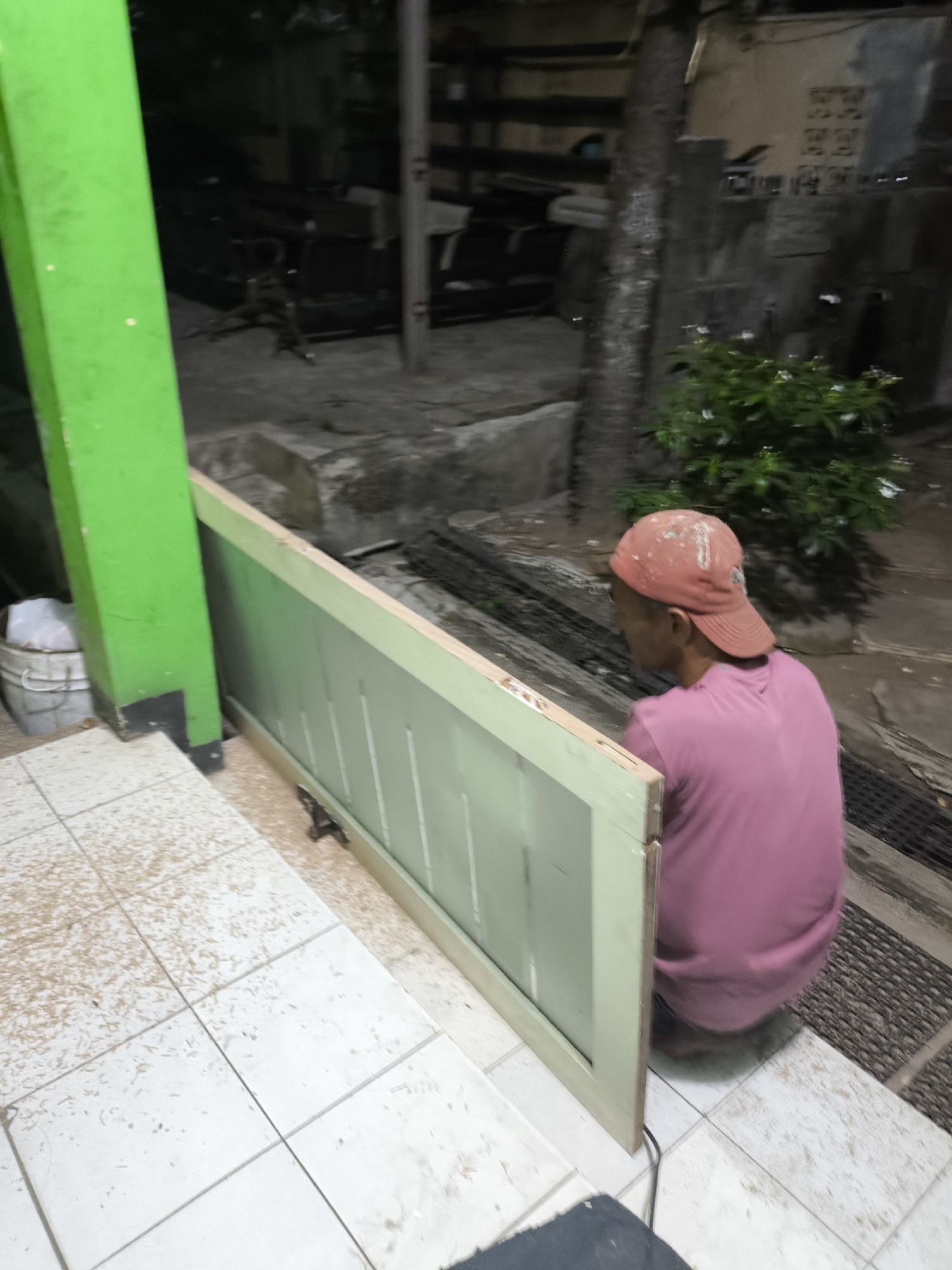 A person wearing a pink t-shirt and a red baseball cap is seen from behind, squatting near a wooden door frame outdoors. The scene appears to be in an urban setting, possibly in Cilegon, Indonesia, given the context. The environment suggests a construction or renovation area, with a green pillar on the left and white tiled floor. The wooden door frame is partially propped against the green pillar, and some debris is visible on the tiles. In the background, there are more structures and vegetation.