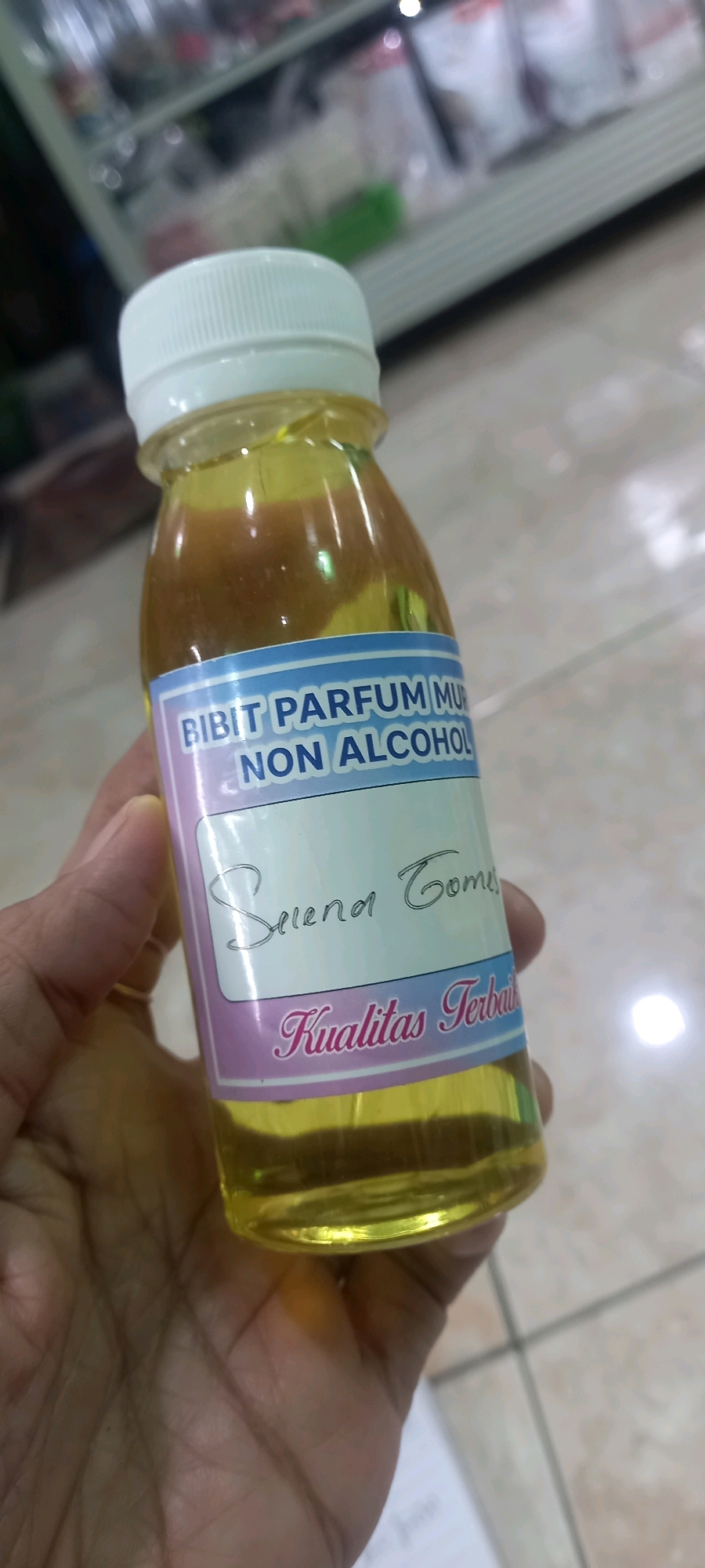 A hand holds a clear plastic bottle containing a yellow liquid. A white cap seals the bottle. A label on the bottle reads "BIBIT PARFUM MU NON ALCOHOL" in blue and white text. Below this, in handwritten script, is "Selena Gomez". At the bottom of the label, in decorative font, it says "Kualitas Terbaik". The background is blurred but appears to be an indoor setting with shelves of goods and a tiled floor. The image was taken in Sedati Agung, Indonesia.