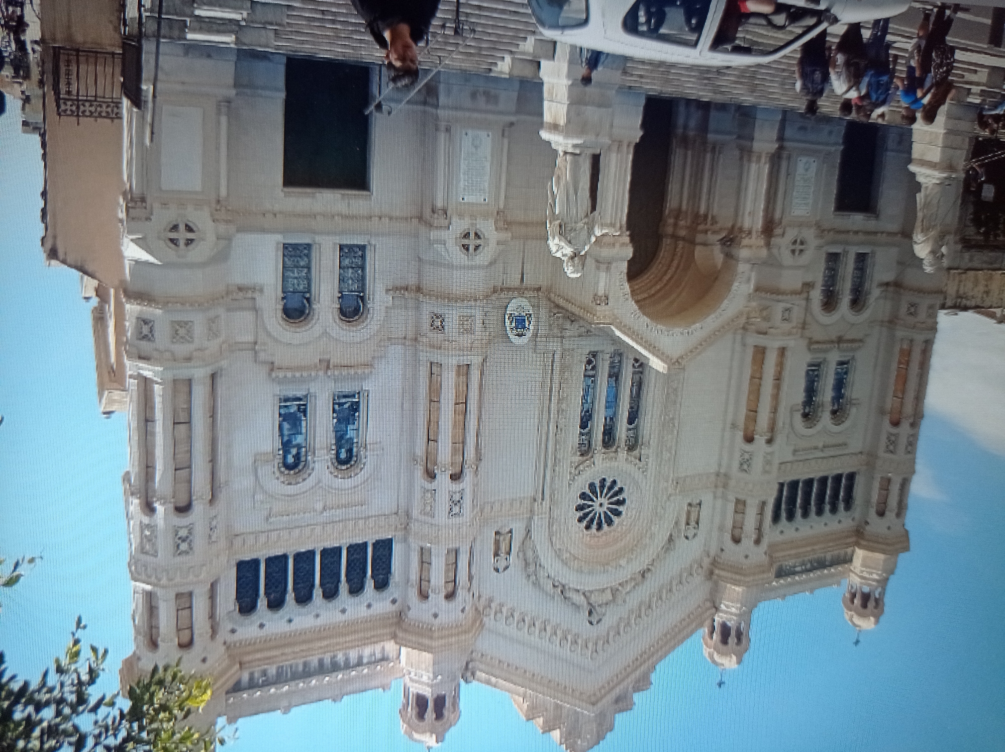 The image shows a grand, ornate white church with intricate architectural details, including arched windows, columns, and decorative carvings. The church has two prominent bell towers topped with crosses. In front of the church, there are wide stone steps leading up to the main entrance. A statue of a robed figure stands on a pedestal to the left of the steps. Several people, including adults and children, are visible on and around the steps, some carrying backpacks. A white car is parked in the foreground, partially obscuring the bottom of the steps. The sky is a clear blue. There are green leaves from a tree visible at the top right corner of the image. The image is taken from a slightly low angle, looking up at the church.
The provided information states this is the city of Mukachevo, Ukraine. The architecture of the building suggests it could be a cathedral or a large church, possibly in a European style. The presence of people and a car indicates it is an accessible public space.