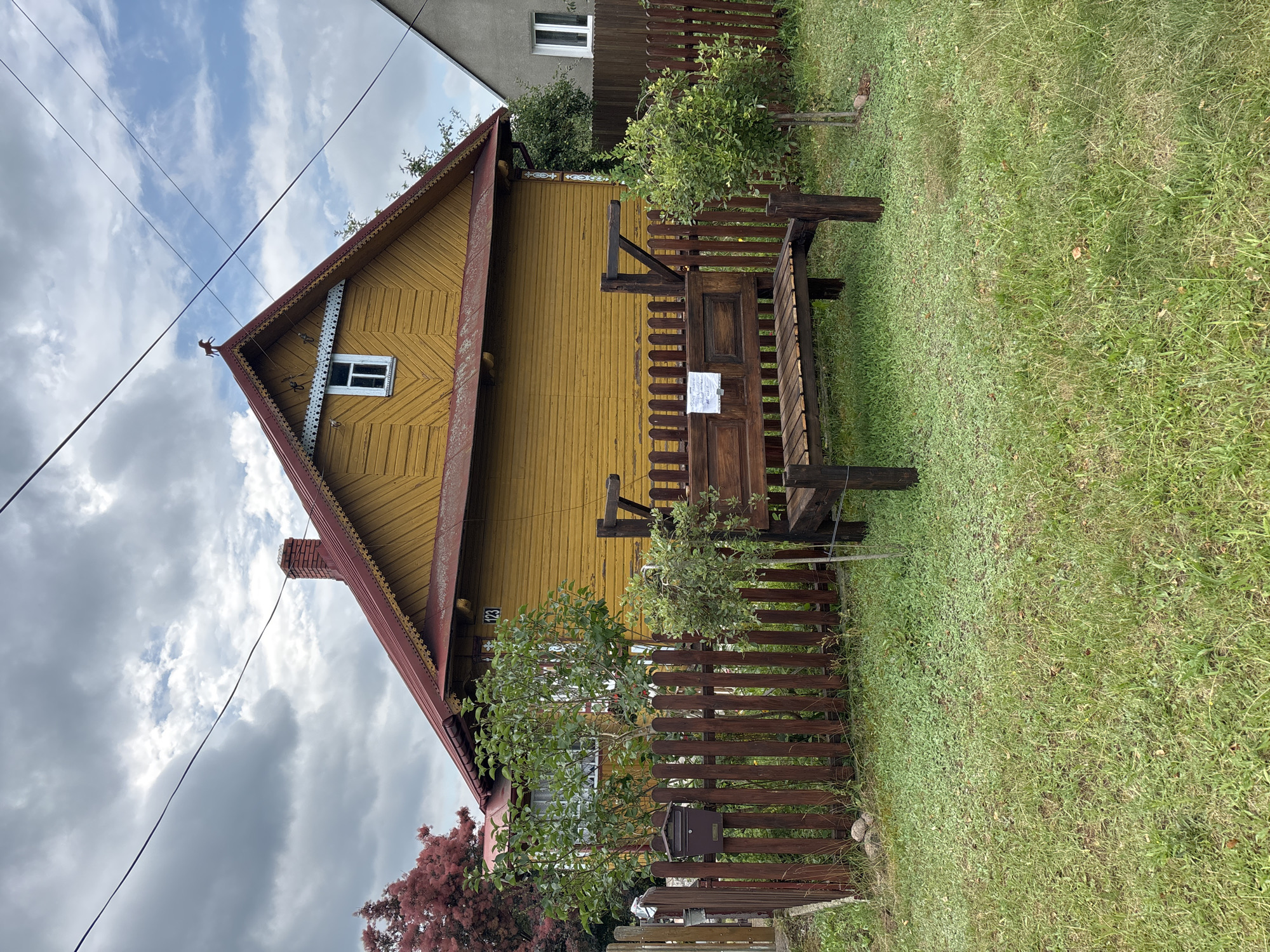 The image captures a scene in the village of Trześcianka, Poland. A weathered, mustard-yellow wooden house with a red roof and decorative eaves is the prominent structure. A red-brick chimney rises from the left side of the roof, and a small, white-paned window is visible on the gable end, topped by a decorative weather vane shaped like a horse. A dark brown wooden fence surrounds a grassy yard in front of the house. In the yard, a rustic dark brown wooden bench sits on the grass. A mailbox is attached to the fence near a leafy green tree. The sky is overcast with dramatic clouds.