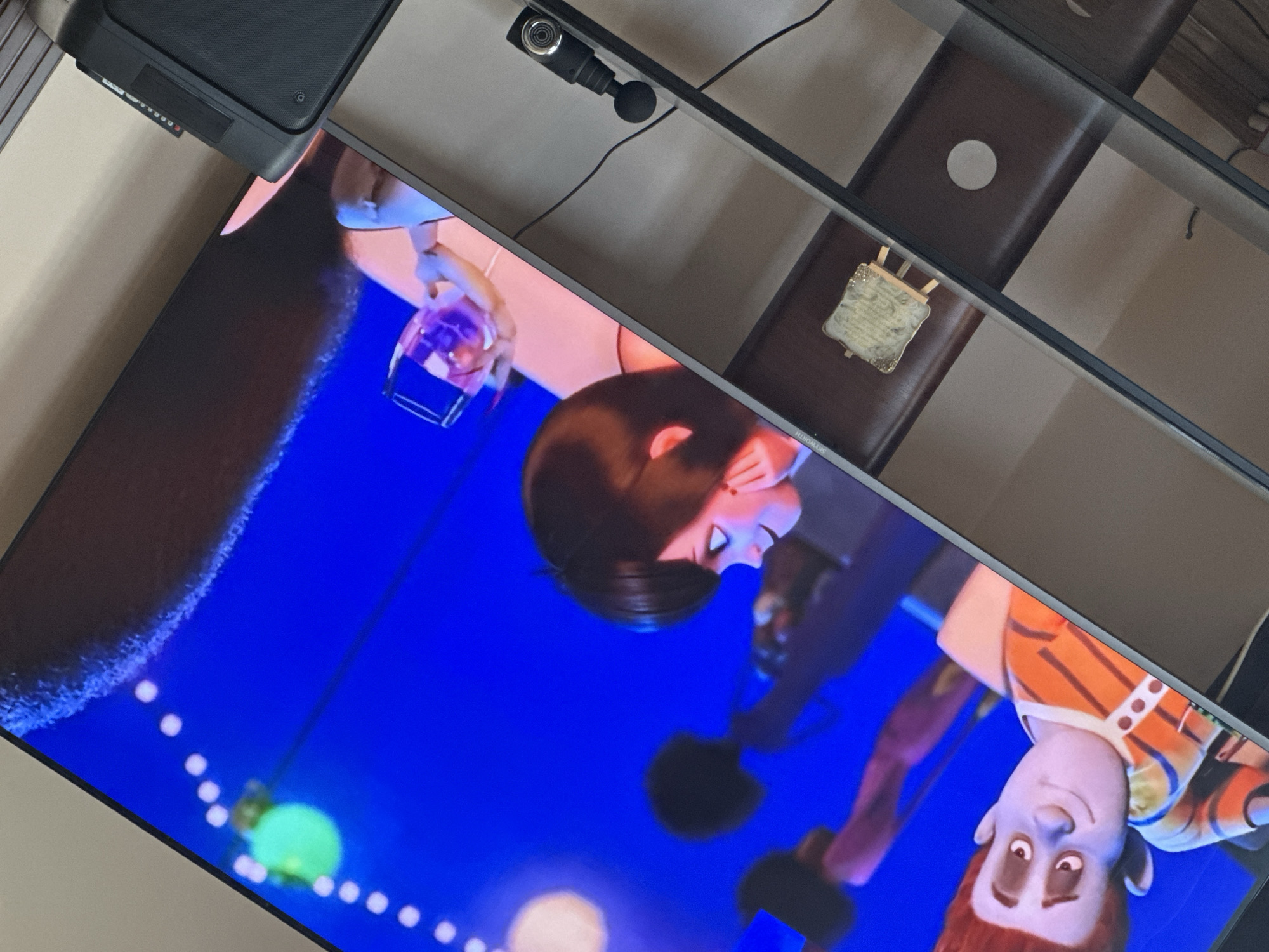 A television screen displays characters from an animated movie. Above the TV is a shelf with a small camera. On the shelf, to the side of the TV, there's a small trophy on a dark wooden surface. The location is indoors, likely a private residence, but the city of Yerevan, Armenia, cannot be determined from the visual data.
