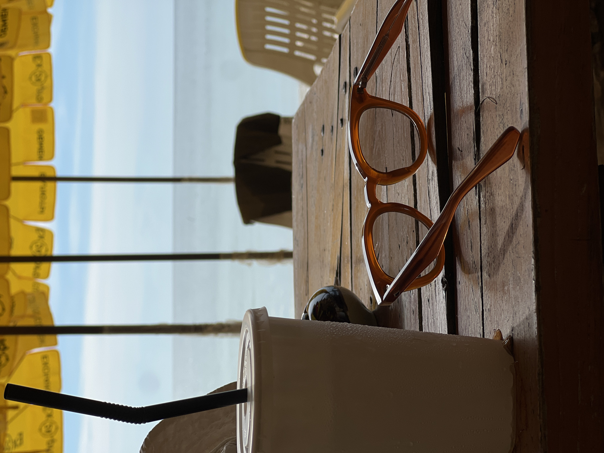 On a wooden plank table, a pair of amber-colored eyeglasses rests next to a white plastic cup with a black straw. A partially visible pair of sunglasses is behind the cup. In the background, yellow umbrellas with writing on them line the sky, which shows through between the umbrella poles. A portion of a white plastic chair is behind the table. The location is the city of Unknown, Unknown.