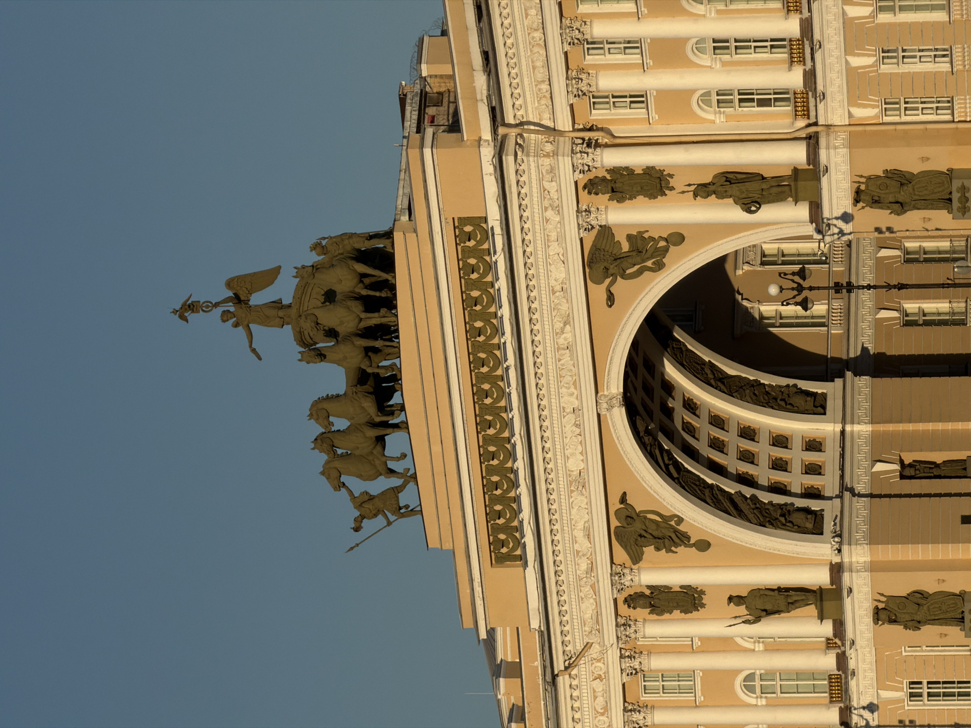 The image shows the Triumphal Arch of the General Staff Building in Palace Square, Saint Petersburg, Russia, bathed in the warm light of either sunrise or sunset, casting long shadows. The arch is adorned with sculptures, most notably the Chariot of Victory at its apex, featuring a goddess holding a laurel wreath and a globe. Below the arch, the building's facade is a pale yellow, with white classical architectural elements like columns, pediments, and decorative friezes. Statues of warriors and allegorical figures are positioned in niches and on pedestals along the facade. The sky above is a clear, bright blue, contrasting with the warm tones of the architecture.