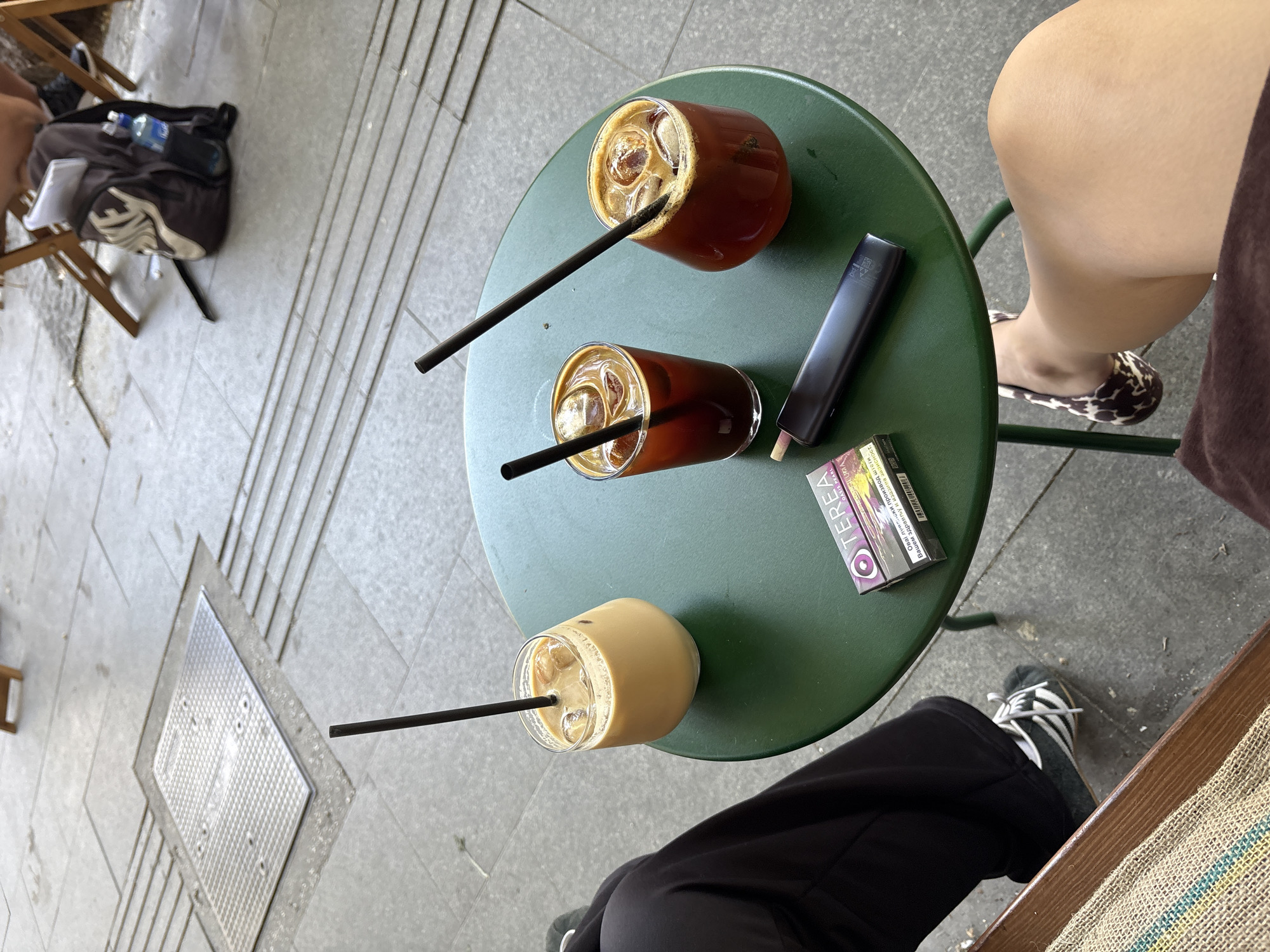 A high-angle shot shows a round, dark green table outdoors on a paved surface. On the table are two iced coffee drinks, a pack of cigarettes, and a dark electronic device. One person's legs, wearing black pants and white-striped sneakers, are visible on the left side of the frame. Another person's legs, wearing a brown skirt and leopard-print flats, are on the right. In the background, a backpack and two wooden chairs are visible. The paving stones in the background suggest a public area, possibly a cafe or restaurant patio in Belgrade, Serbia.