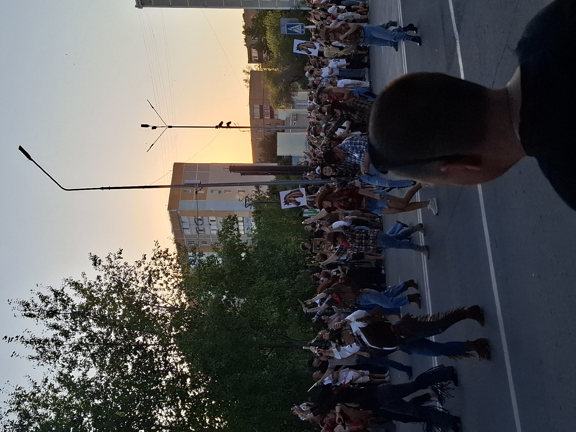 A group of people in cowboy attire are marching in a street, with some holding up signs with horse images. The sun is setting, casting a warm glow on the scene. Buildings line the street, and trees are visible in the background. The people are dressed in jeans, cowboy hats, and boots, with some wearing vests and bandanas. They appear to be participating in a parade or festival. The overall atmosphere is festive and lively.