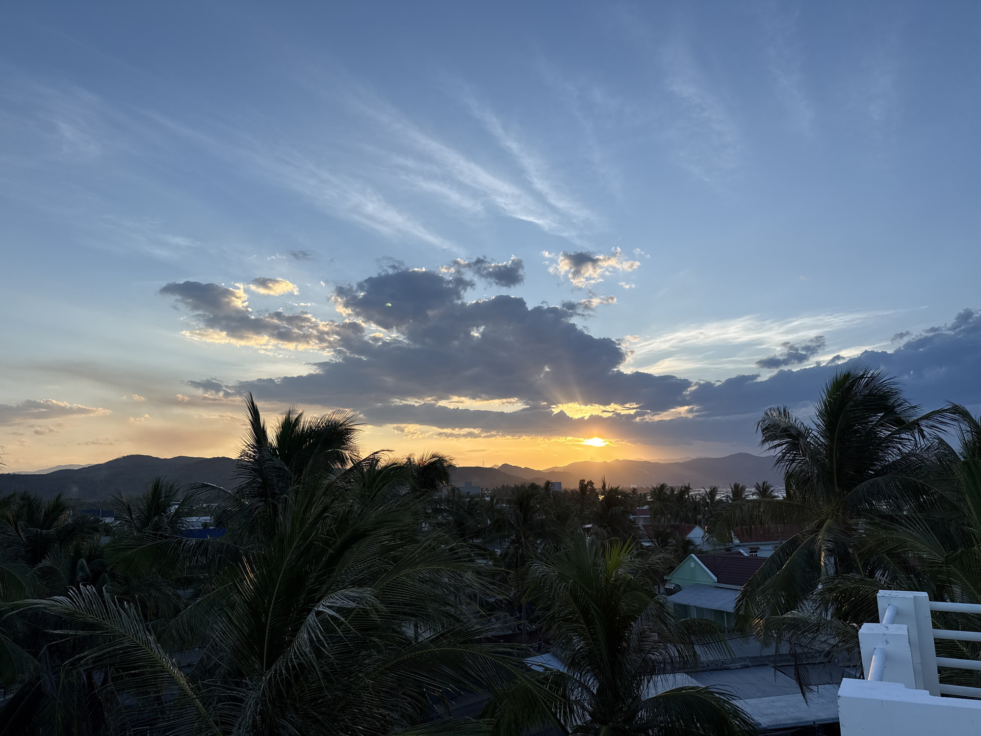 A wide-angle shot captures a serene sunset over a tropical landscape. The sky is a canvas of soft blues and oranges, with wispy clouds streaking across the upper half of the frame. The sun, a radiant golden orb, peeks through a gap in the clouds, casting long, ethereal rays of light downwards. In the foreground, lush palm trees dominate the scene, their fronds silhouetted against the vibrant sky. Below, a small town with buildings and rooftops is nestled amidst the greenery, with a range of mountains forming a distant backdrop. The overall mood is peaceful and tranquil, evoking a sense of warmth and serenity.