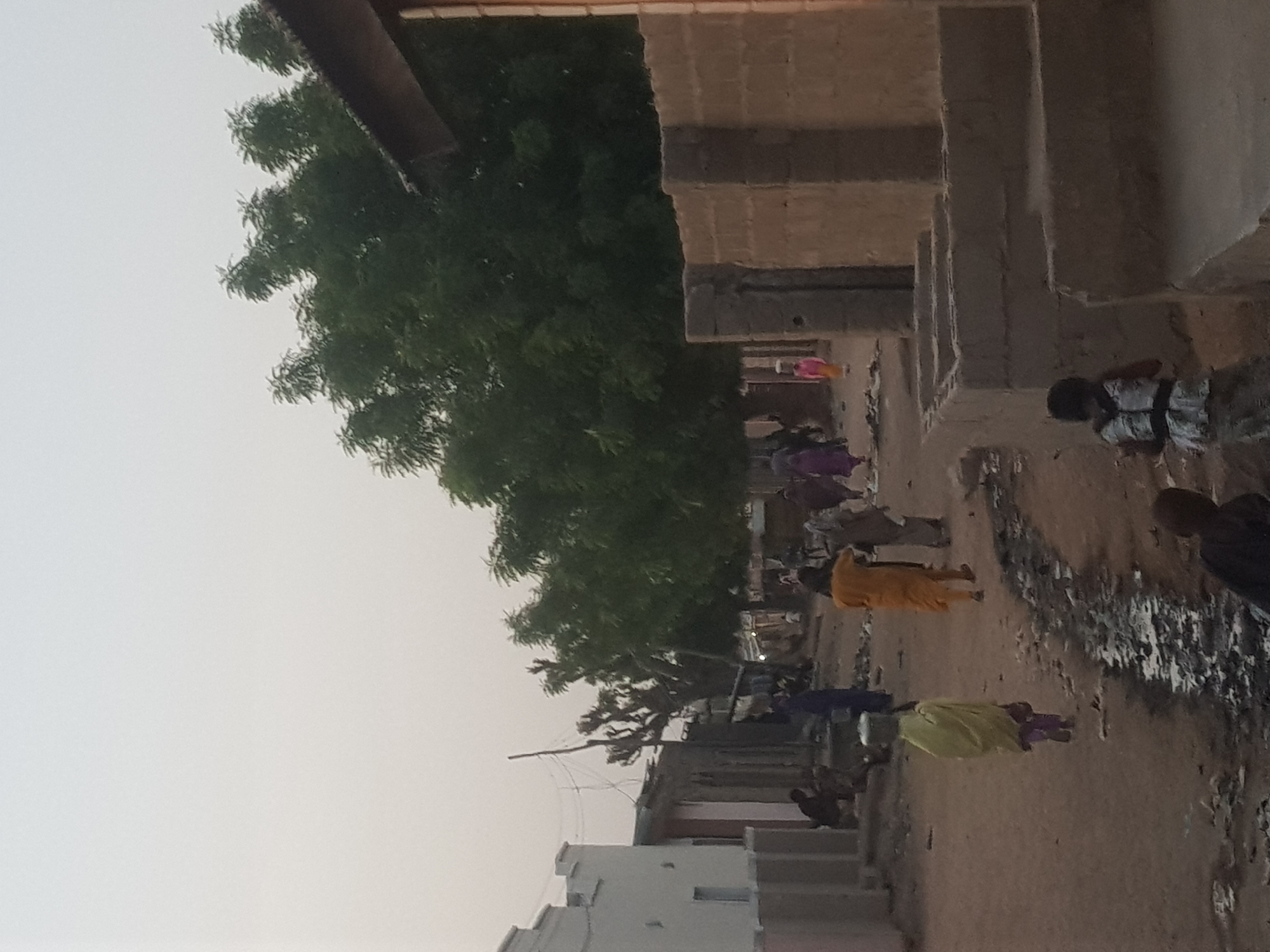 The image depicts an unpaved street scene in Dutsinma B, Nigeria, with multiple people walking along the street. Buildings in various stages of construction line one side of the street. A large, mature tree is also present. The scene appears to be in daylight, with a slightly hazy or overcast sky. There are puddles and debris along the side of the street. The people are wearing a variety of clothing, with some wearing brightly colored outfits.