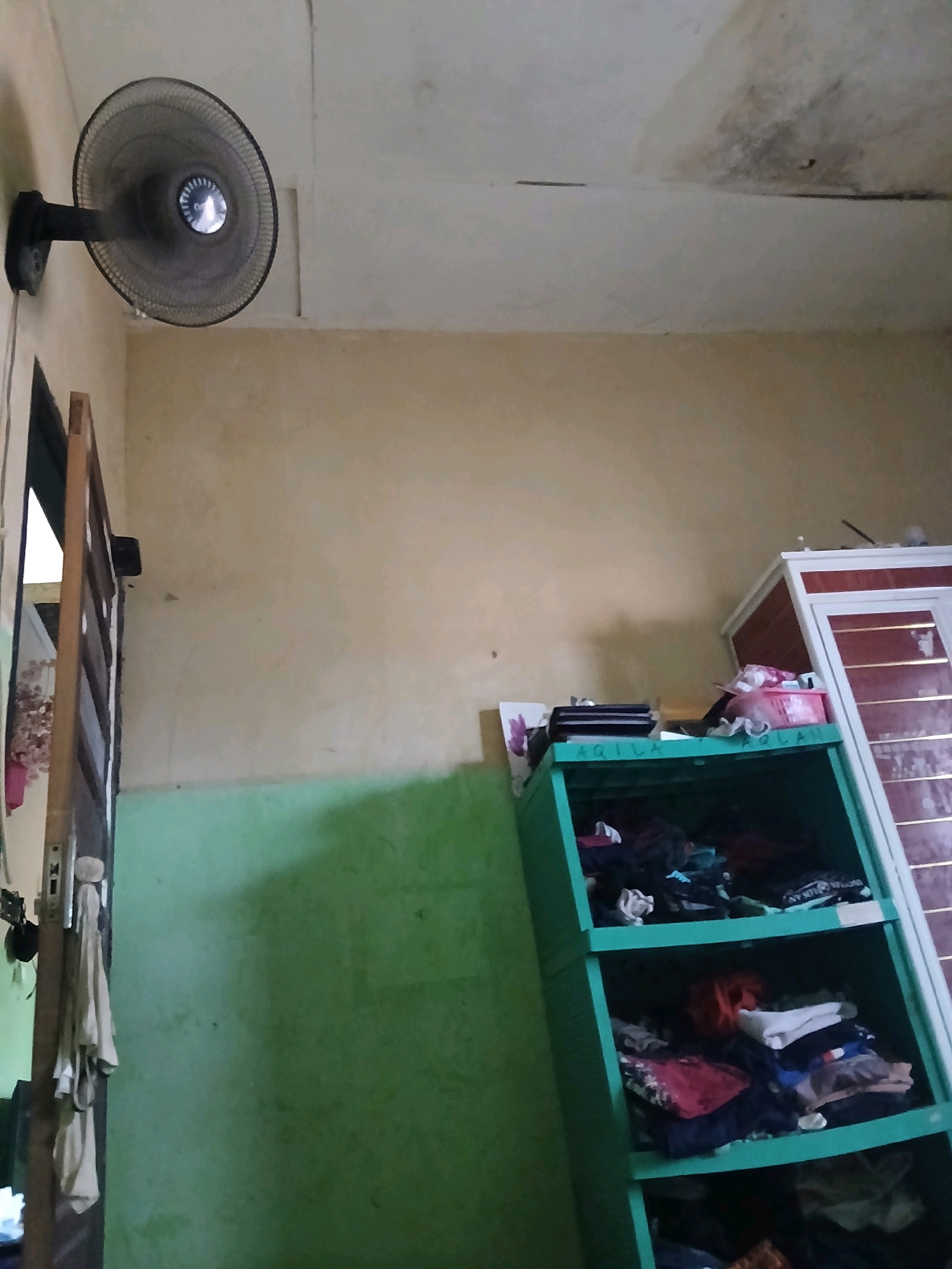 The image depicts an interior scene, likely a residential room, in Pangkalpinang, Indonesia. A black oscillating fan is mounted on a light-colored wall, angled downwards. To the left, a portion of a wooden door frame is visible, with some fabric items hanging on it. The walls are painted in muted tones, with a distinct line separating a light beige upper section from a bright green lower section. In the background, a green plastic storage shelf unit is filled with various folded clothes. Adjacent to this shelf is a taller, white cabinet with reddish-brown accents. The ceiling appears to be discolored in areas, possibly due to water stains. The overall impression is of a utilitarian and lived-in space.